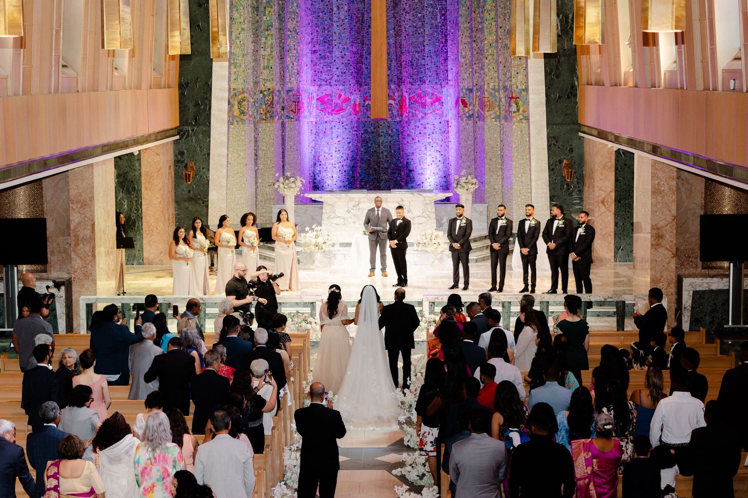 Wedding ceremony in an elegant church with purple uplighting and rows of guests watching the couple exchange vows.