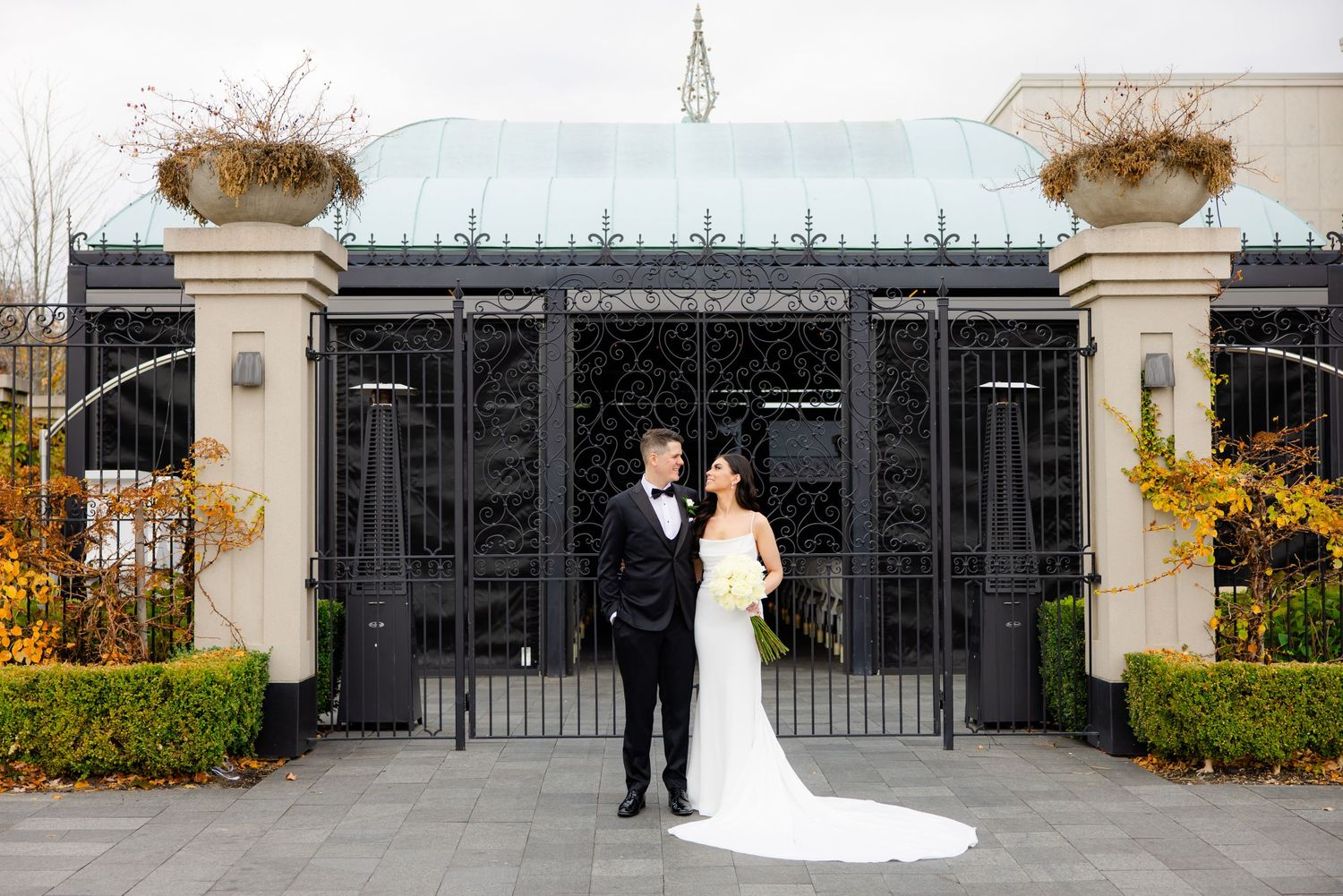 Wedding couple stands before grand entrance gates with ornamental pillars and autumn foliage.