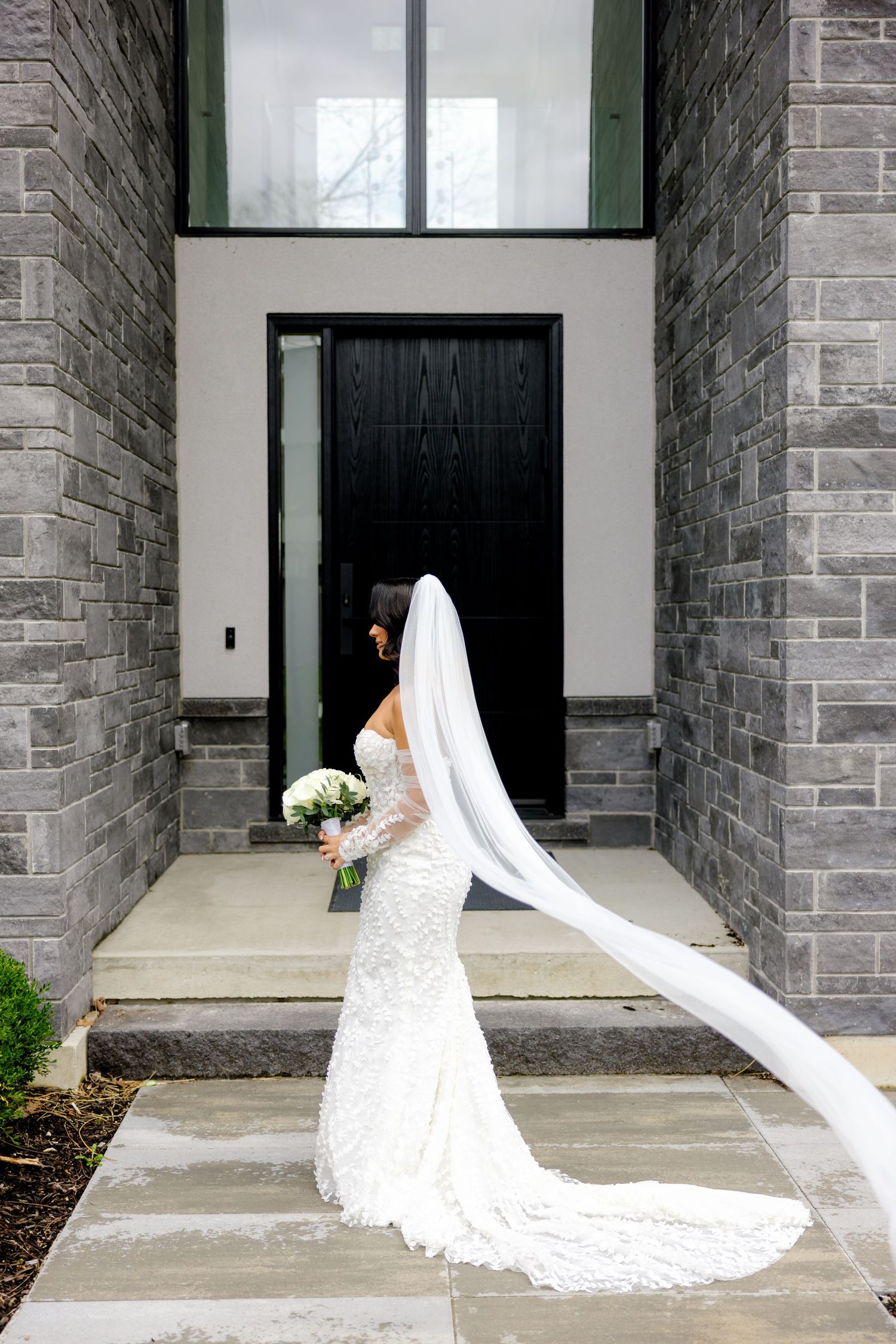 Bride in lace dress with cathedral length veil stands before black door and stone walls.