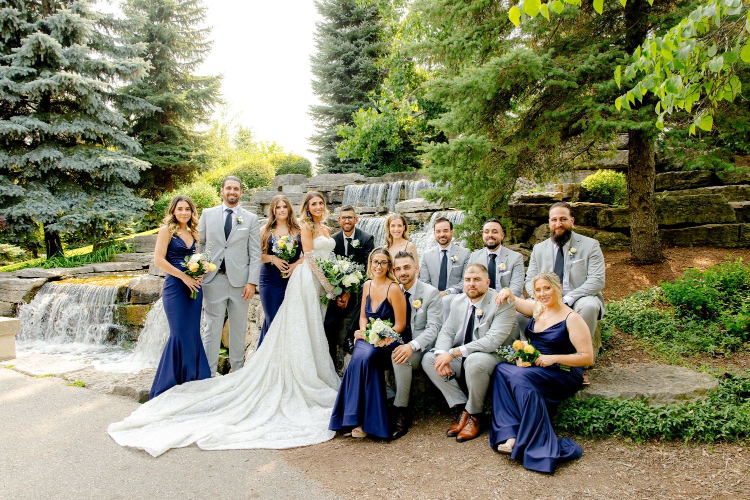 Wedding party in navy and gray attire poses together by garden waterfall.