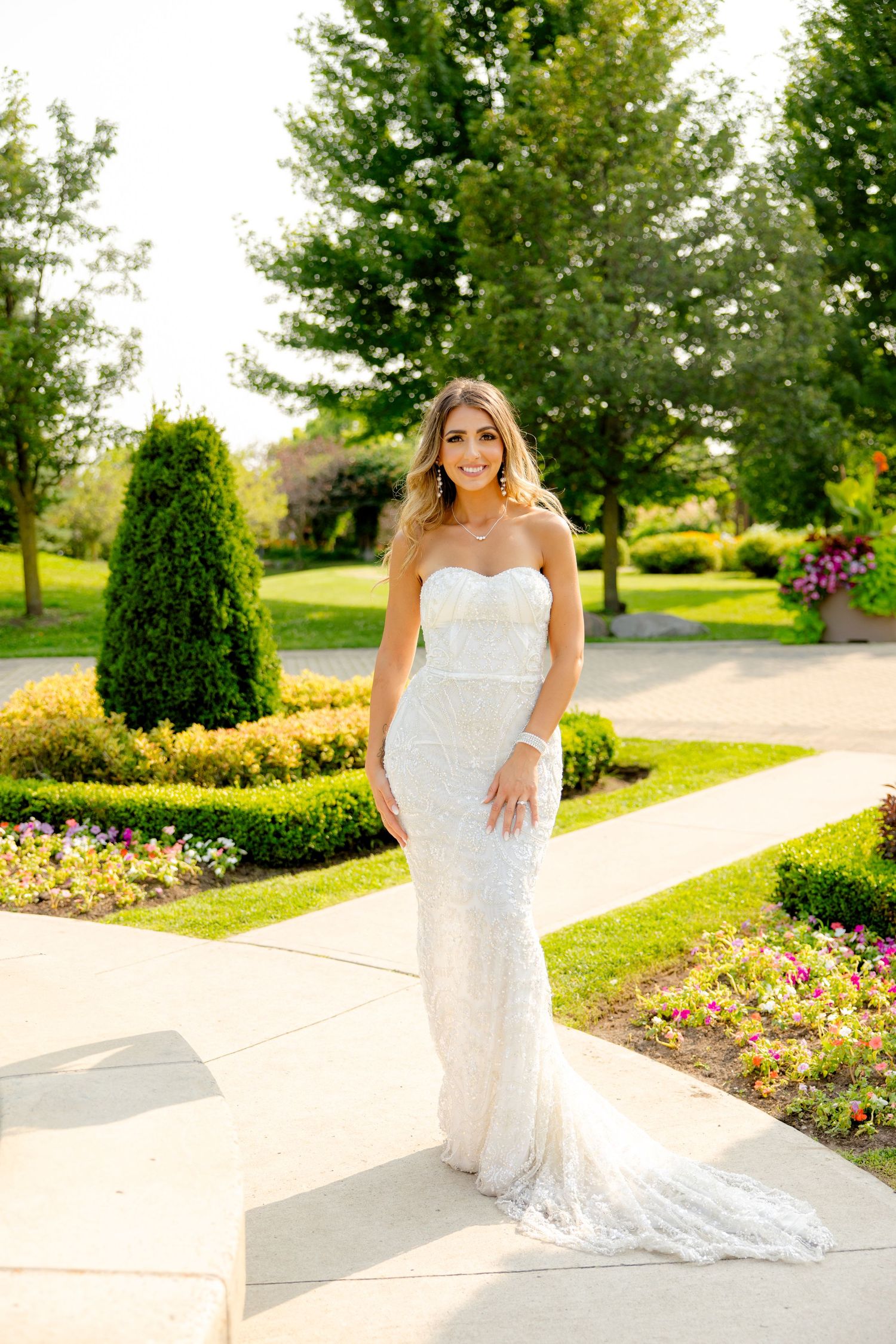 Bride in strapless lace gown poses on garden path with perfectly manicured landscape.