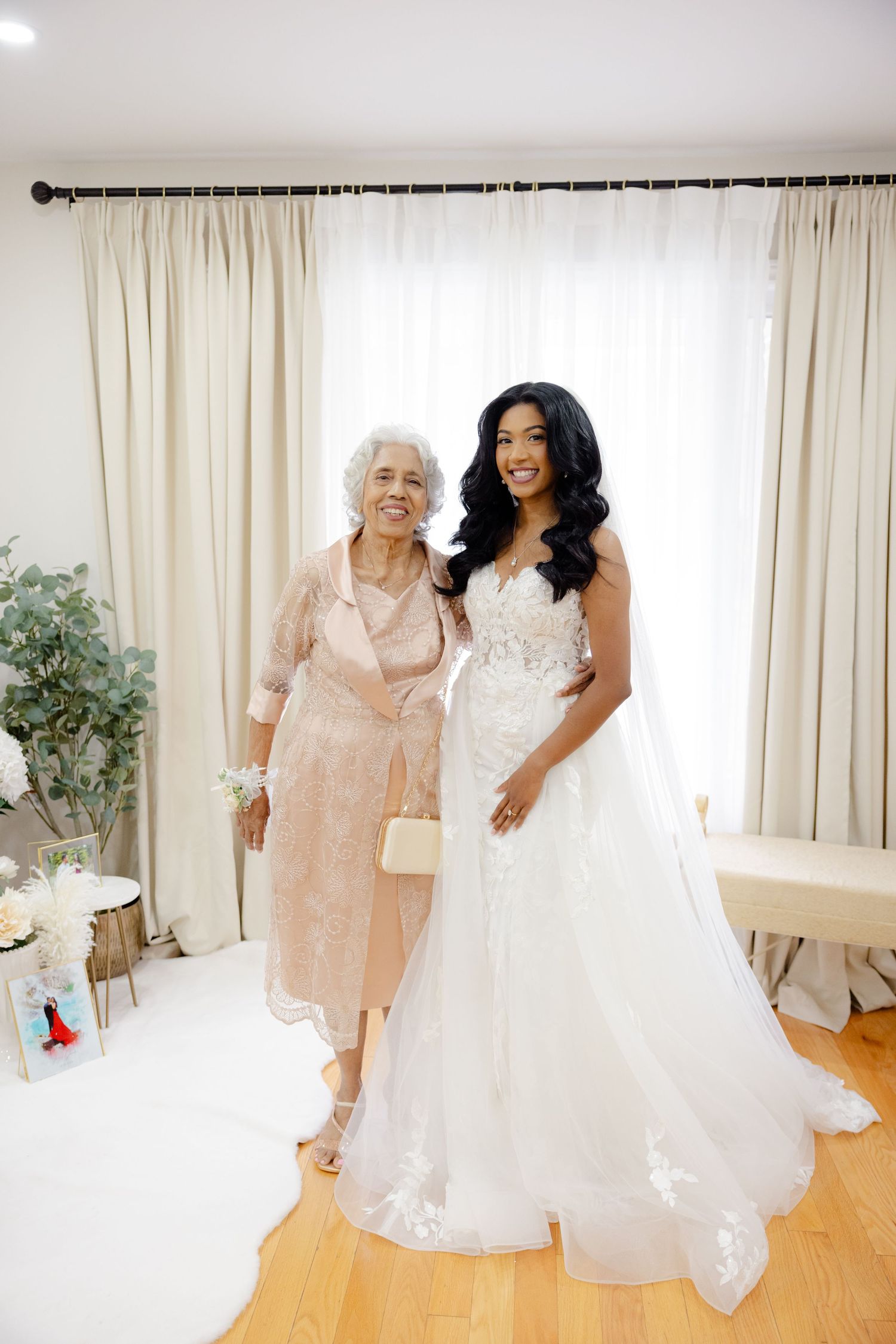 Full-length portrait of bride and guest posing together against light window backdrop.