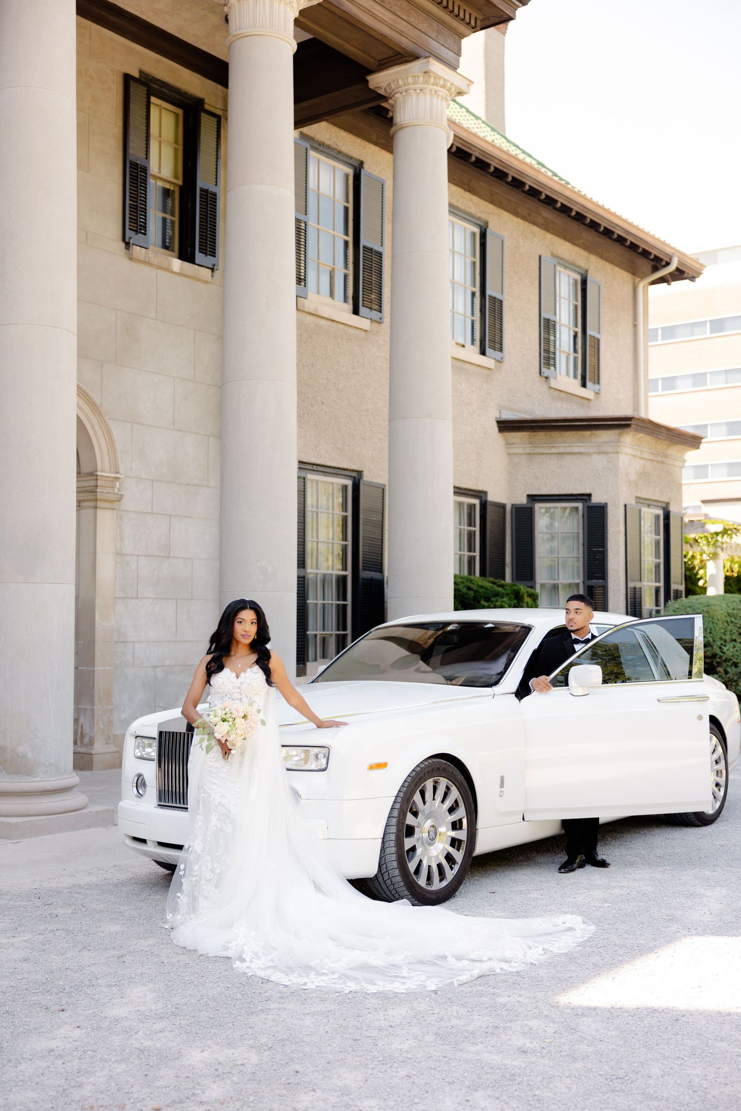Bride and groom stand beside a white Rolls-Royce luxury car at their wedding venue.