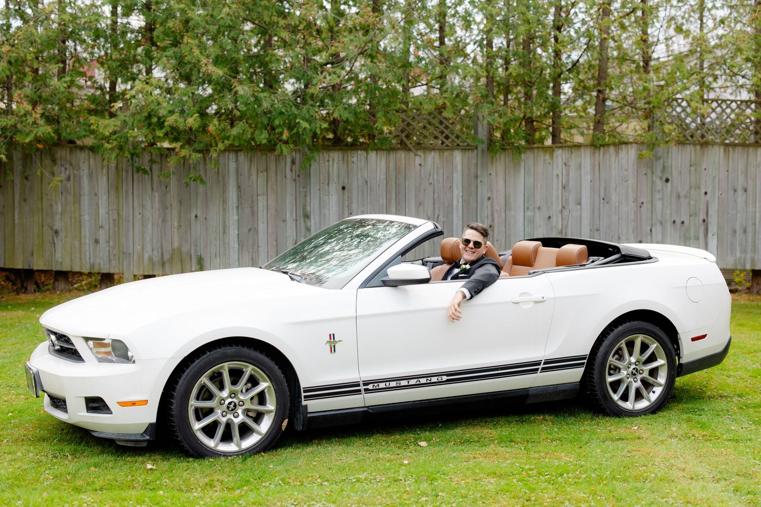 A white Ford Mustang convertible parked on grass in front of a wooden fence.