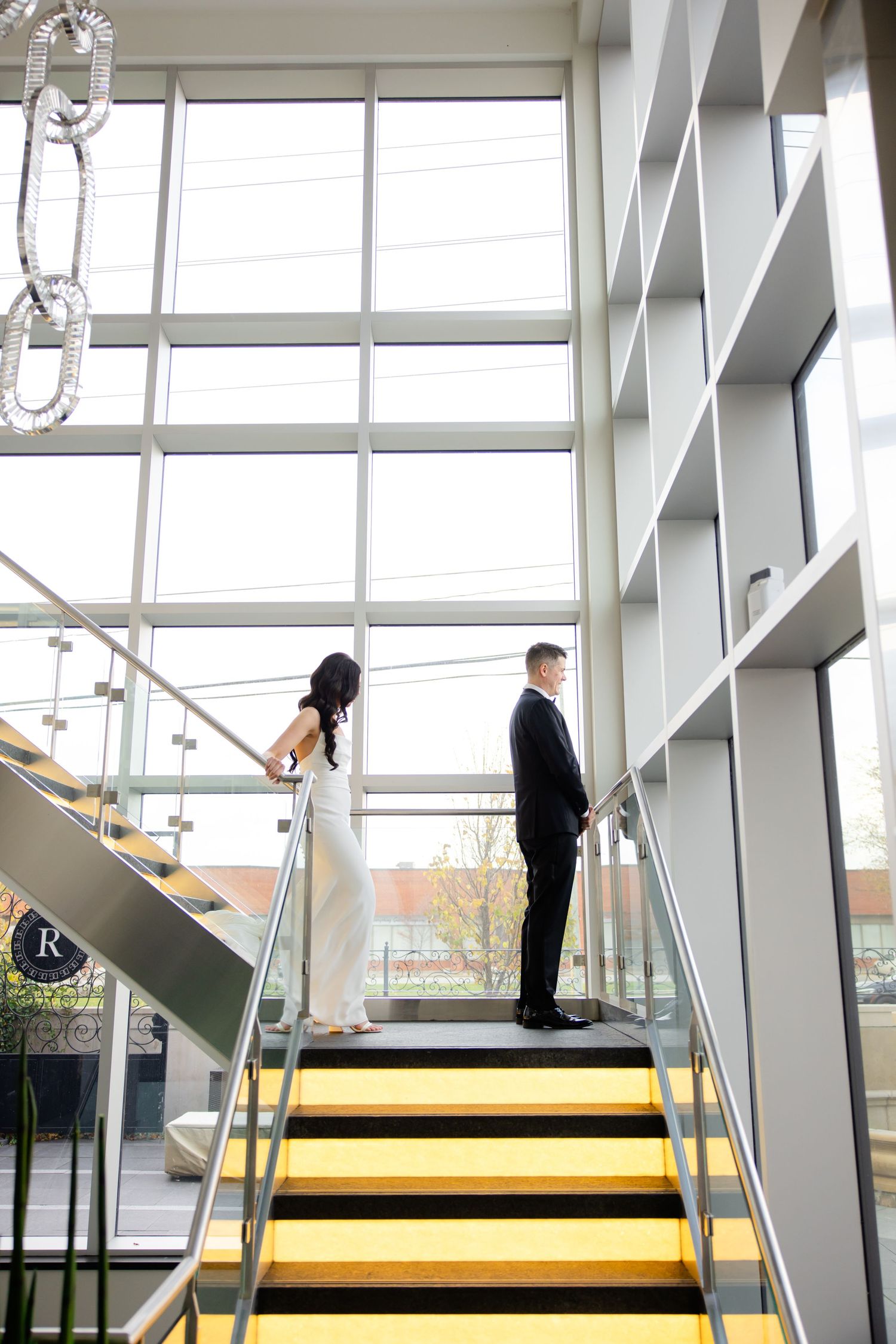 Wedding couple stands apart on yellow-striped stairs in modern building with glass walls.
