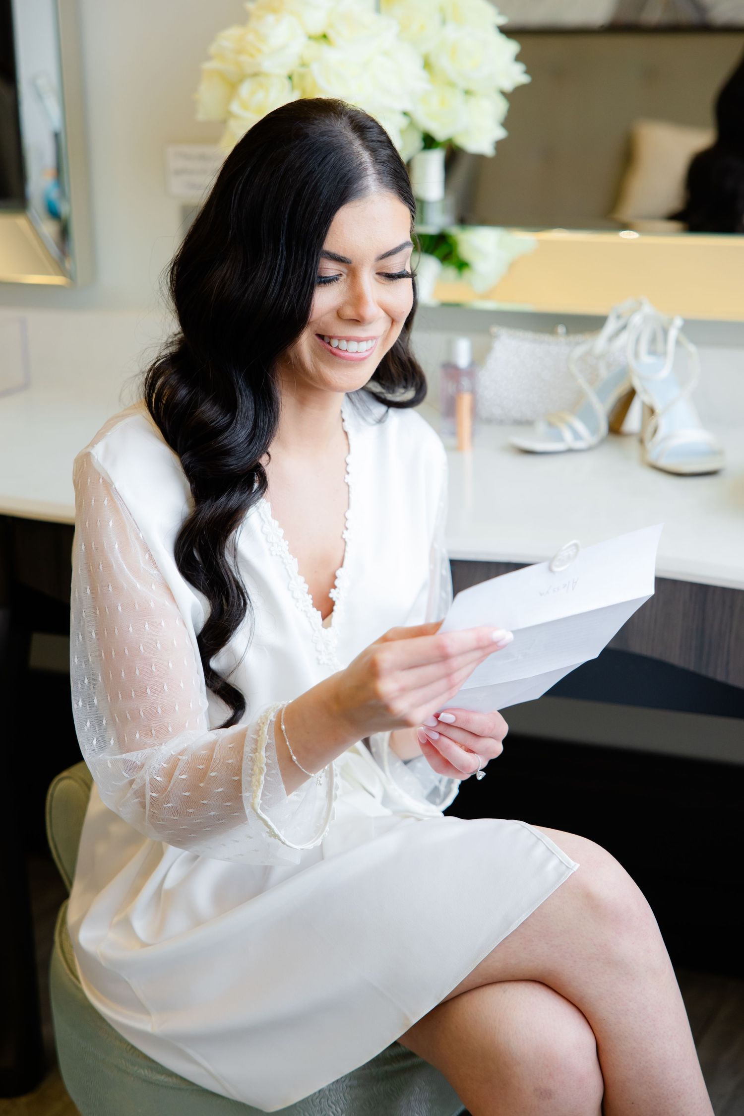 Person in white robe reading letter at vanity with flowers.