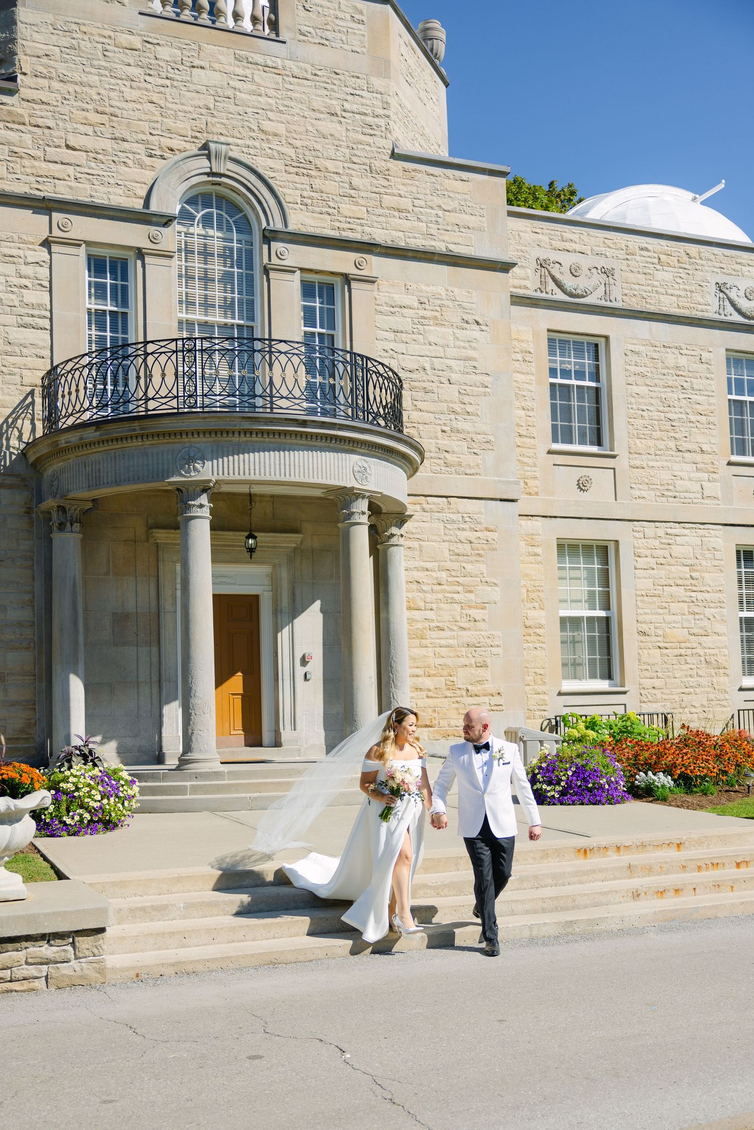 Newlyweds walk down steps of a grand historic building with classical architecture and flower gardens.