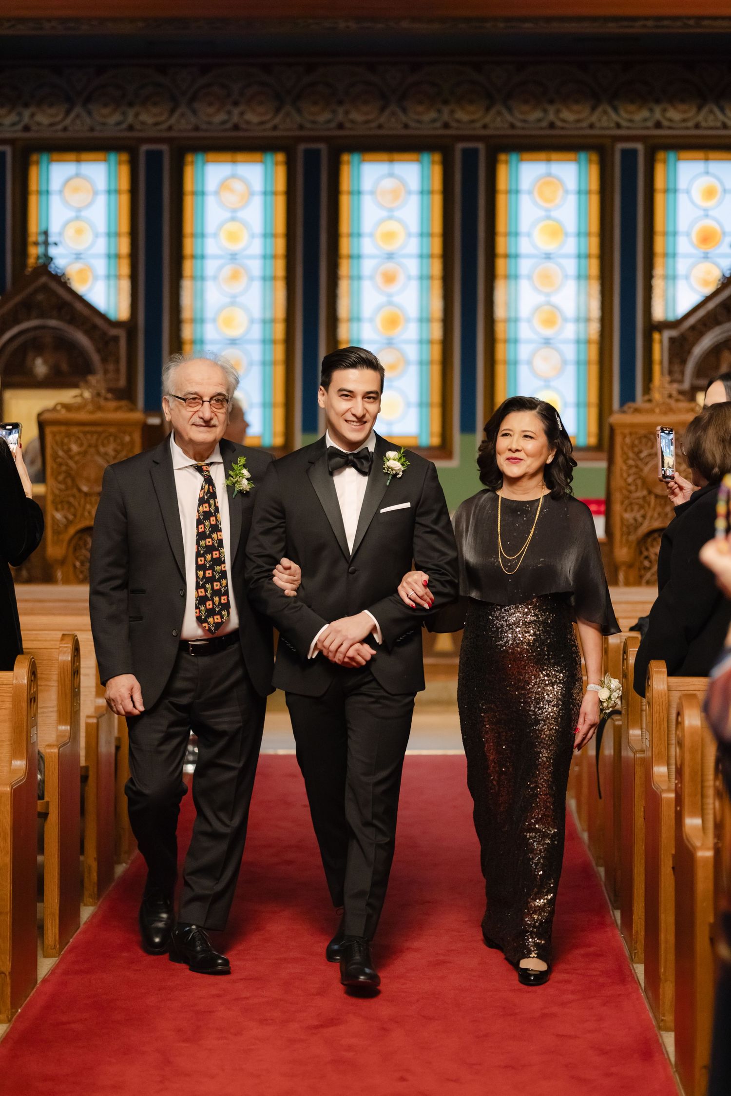 Wedding party members walk down a church aisle with stained glass windows and wooden pews.