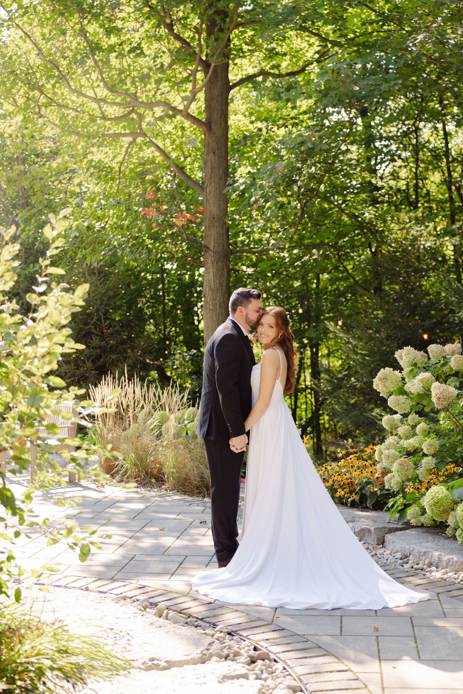 A romantic wedding portrait on a stone path with sunlit trees and white hydrangeas in the background.