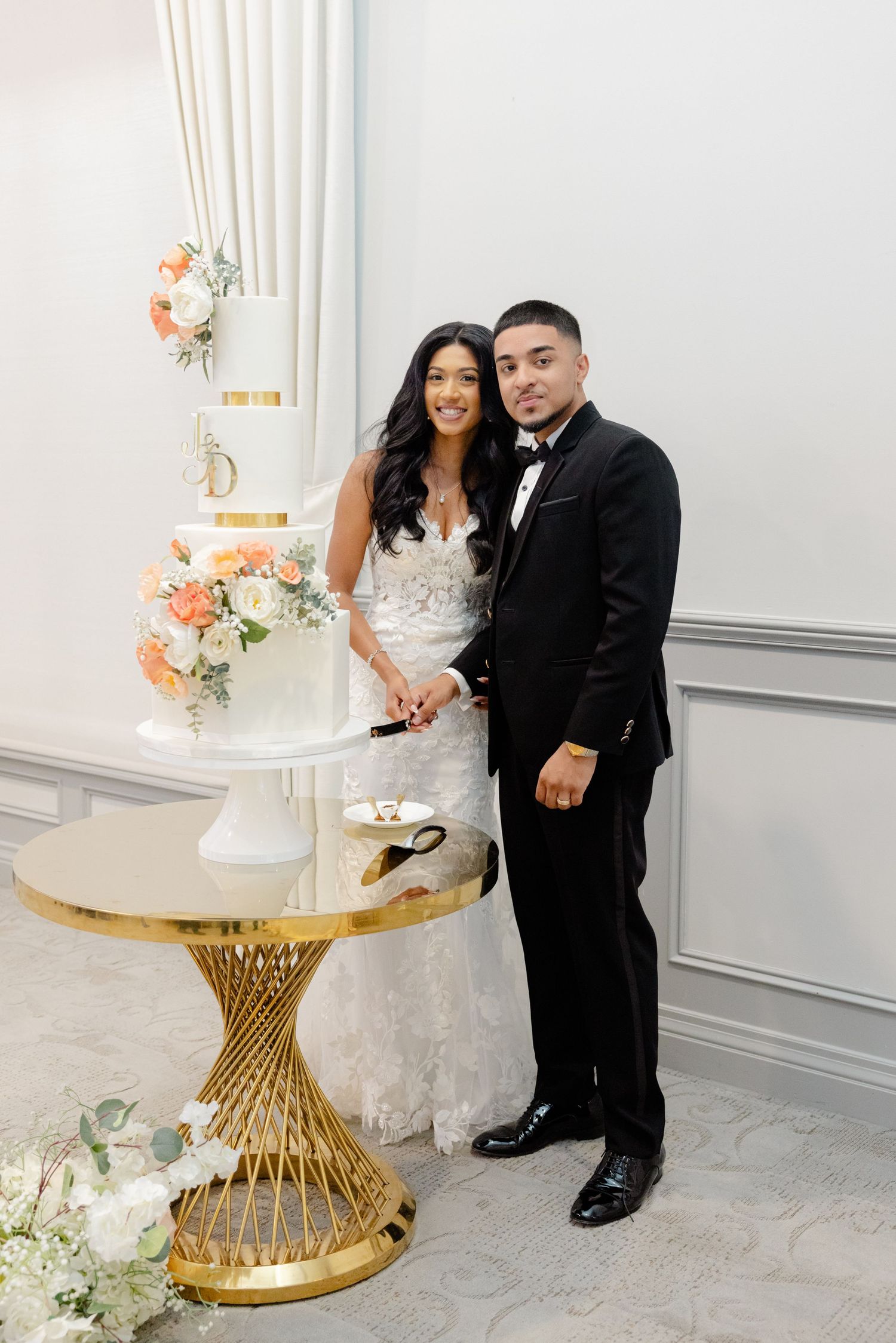 A couple poses together next to their elegant white wedding cake with peach floral decorations.