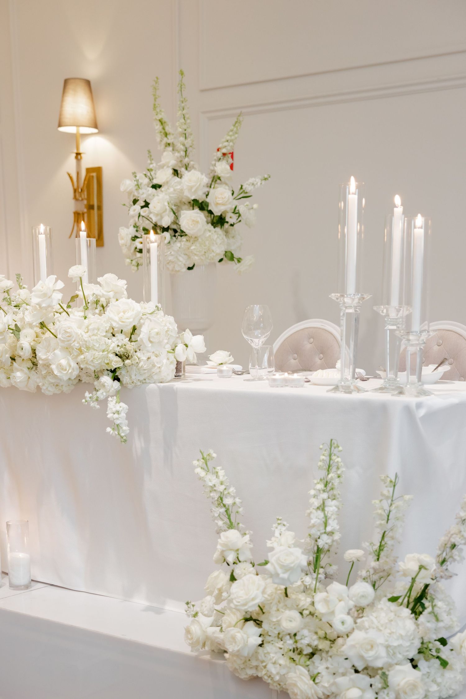 Elegant wedding table setup with white floral arrangements, tall candles, and a white tablecloth in soft lighting.