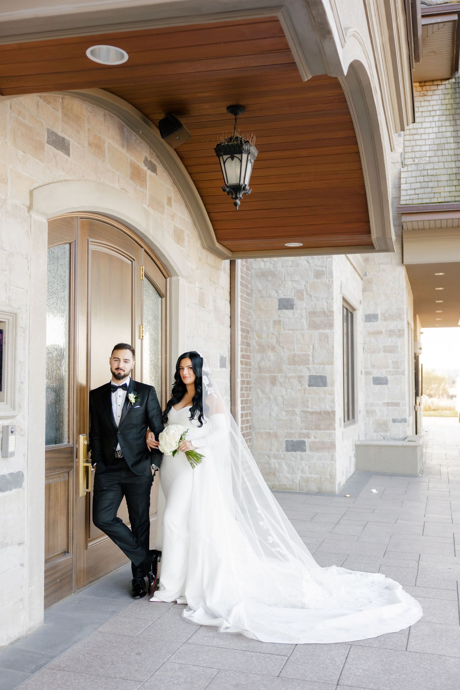 Newlywed couple poses under wooden archway with stone facade and hanging lantern.