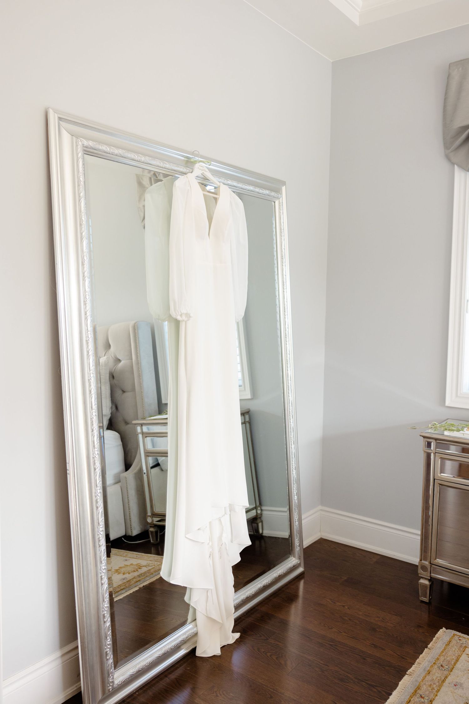 Bridal gown hanging on decorative mirror in modern bedroom with hardwood floors.