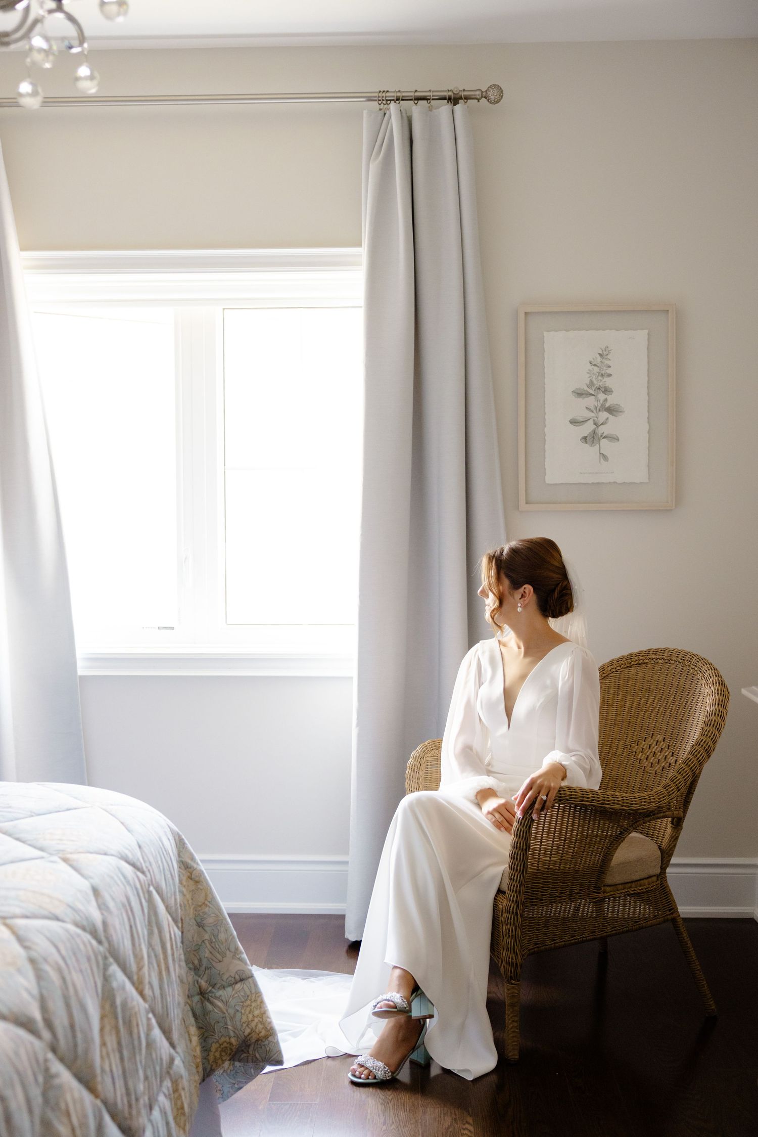 Elegant bridal portrait in a sunlit bedroom with wicker chair and botanical artwork.
