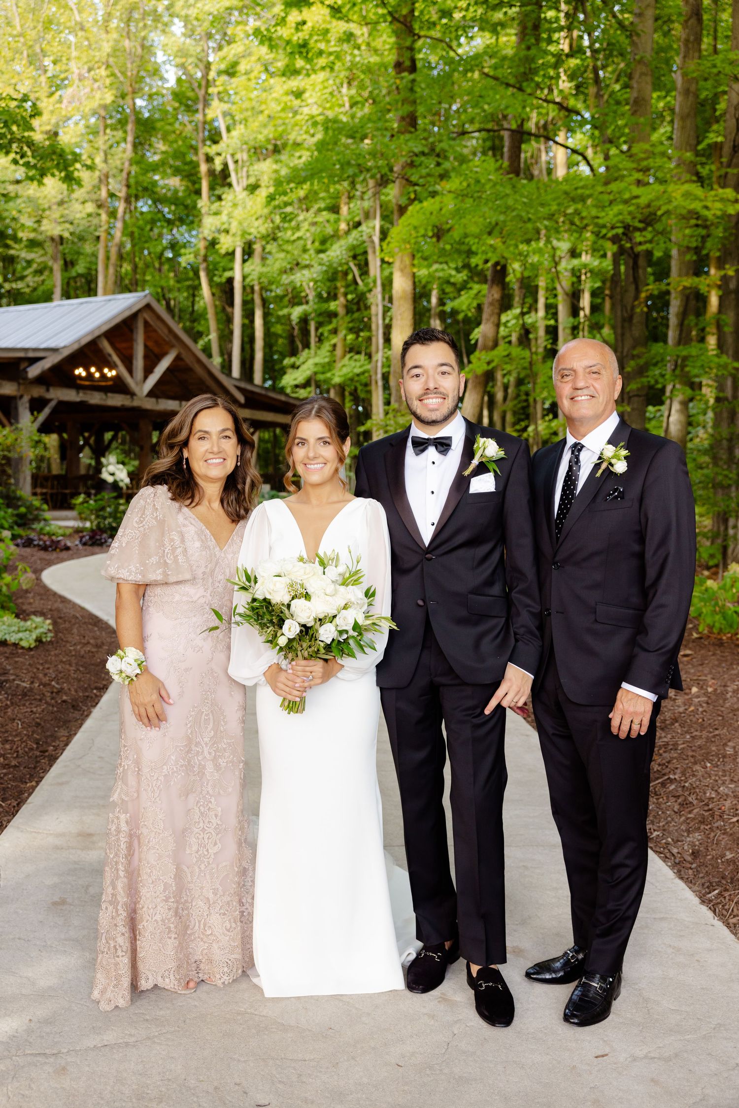 Wedding party poses on concrete path with rustic wooden pavilion and forest backdrop.