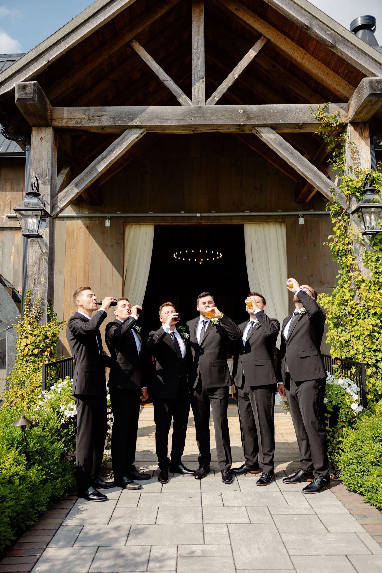 Wedding party in black suits stand together laughing under a rustic barn entrance.