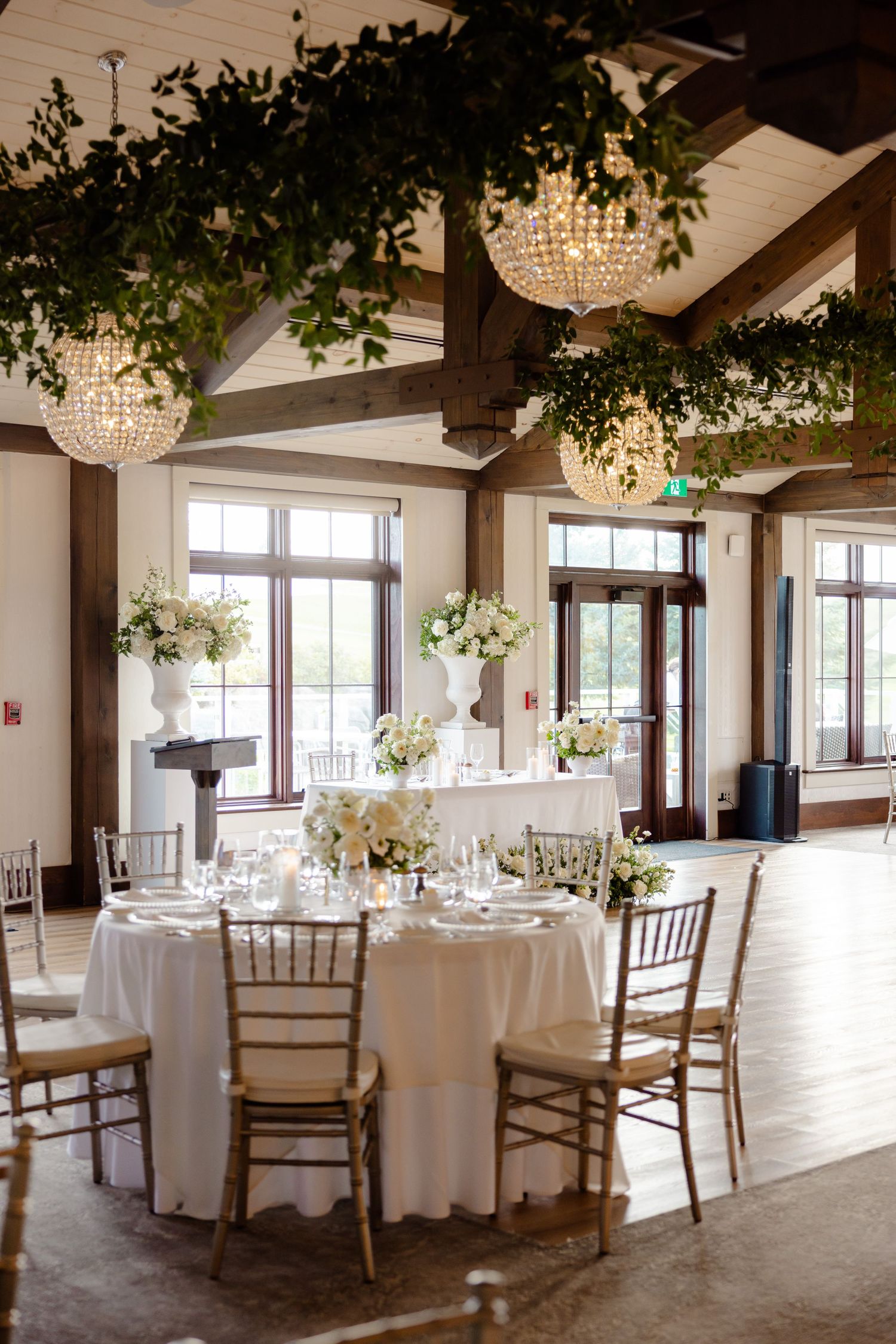 Reception venue with round tables, crystal chandeliers, and greenery-adorned wooden beam ceiling.