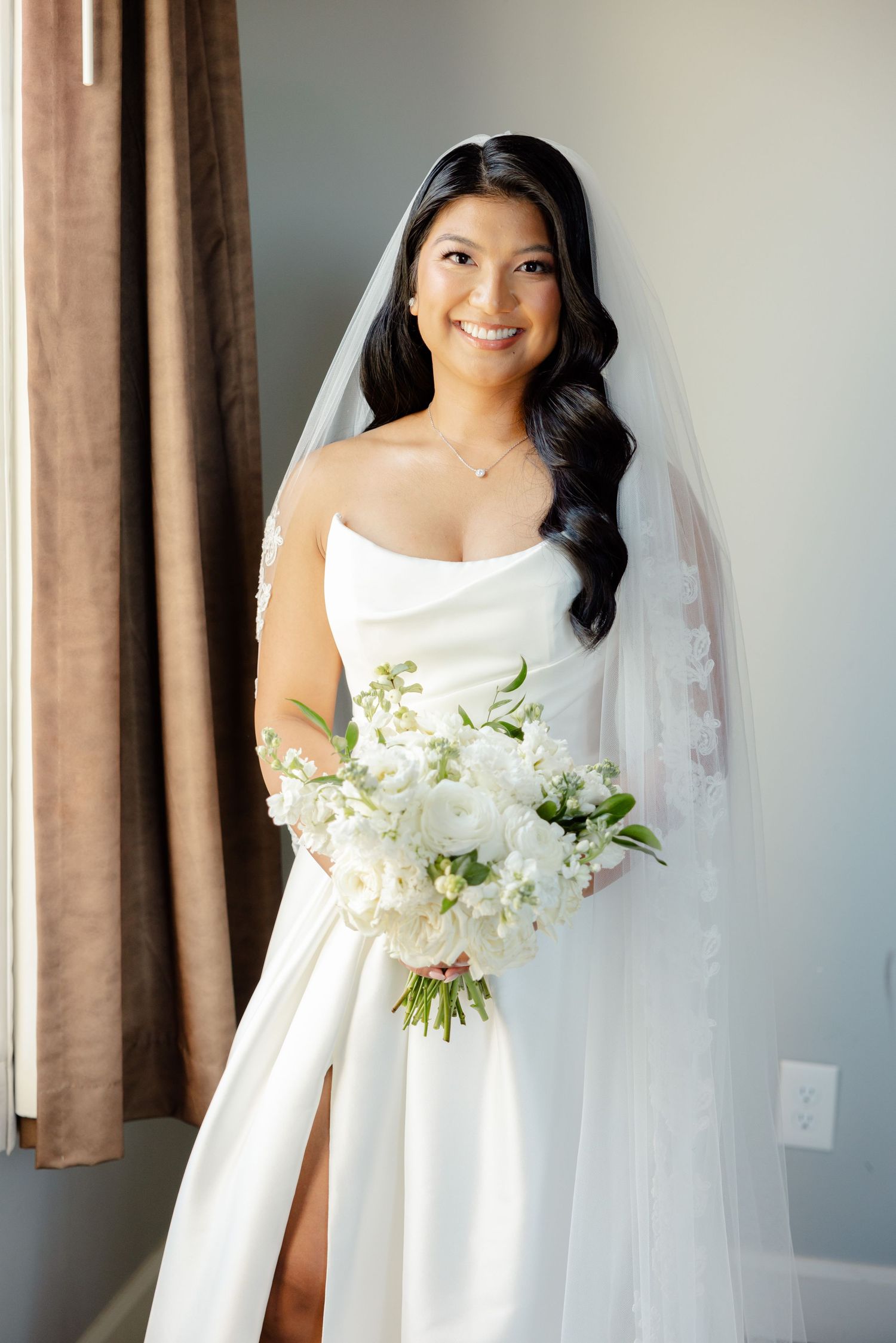 Bride in strapless wedding gown smiles while holding white floral bouquet against neutral background.