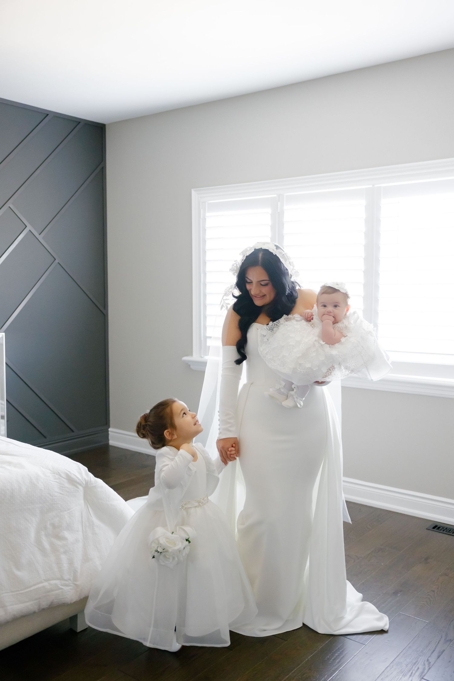 A bride holds hands with flower girls in matching white dresses against a modern gray accent wall.