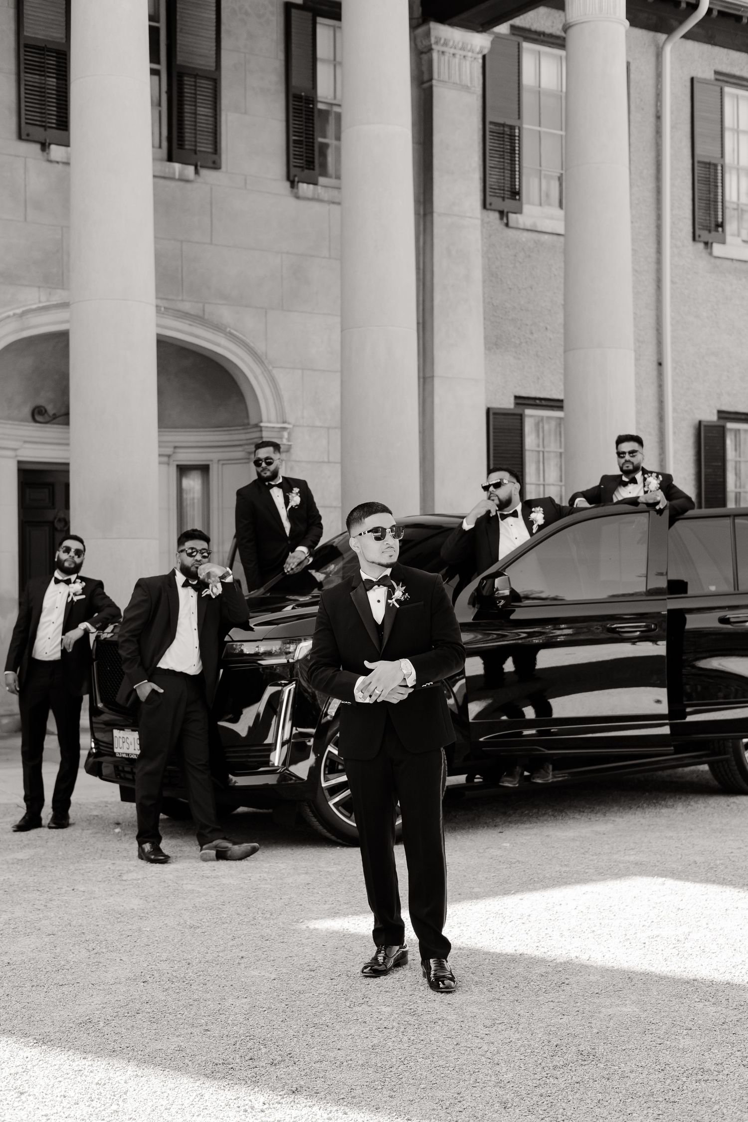 Formal wedding party members strike dynamic poses with a black luxury car at an elegant venue.