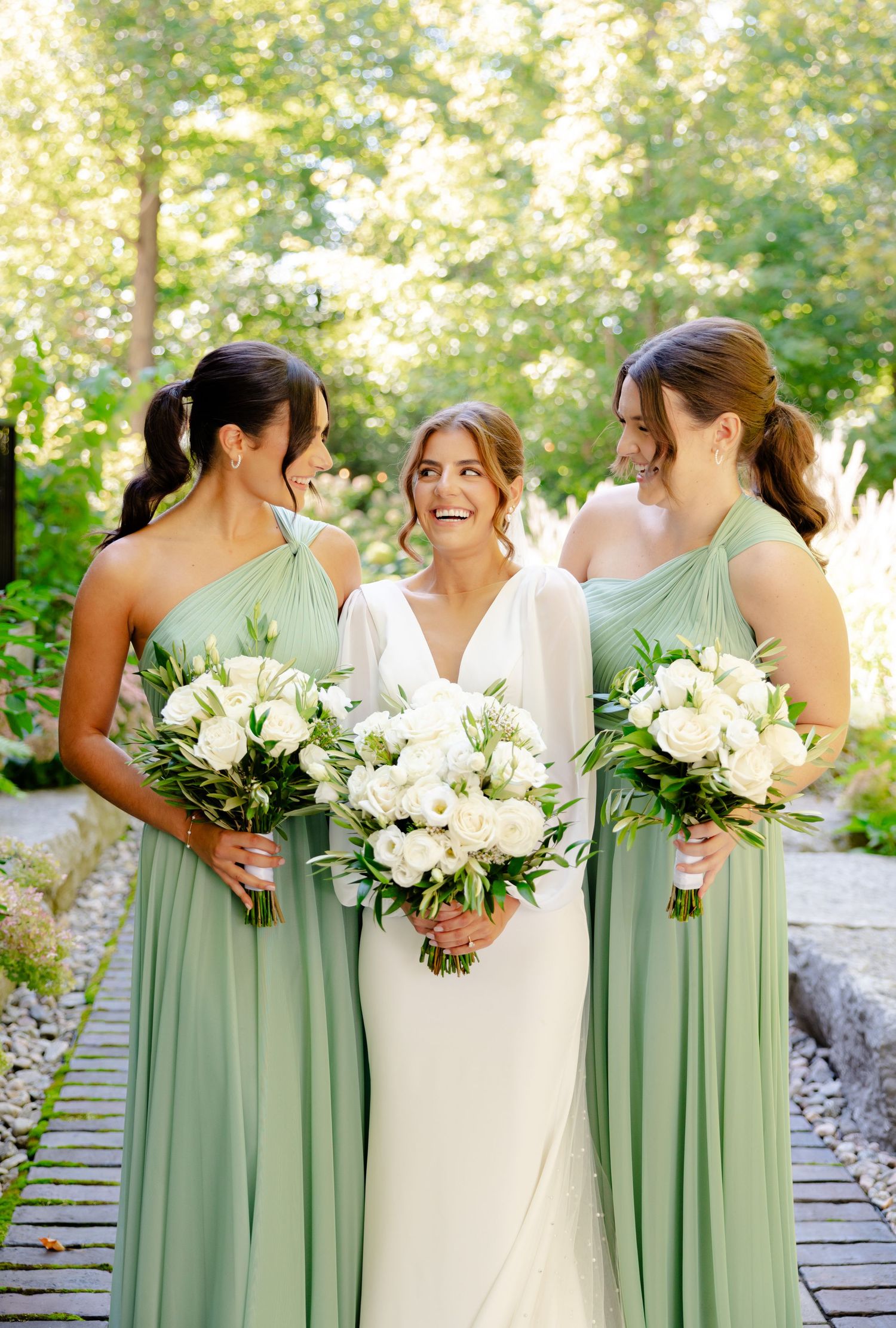 Bridesmaids in mint green dresses share joyful moment with bride holding white bouquets outdoors.