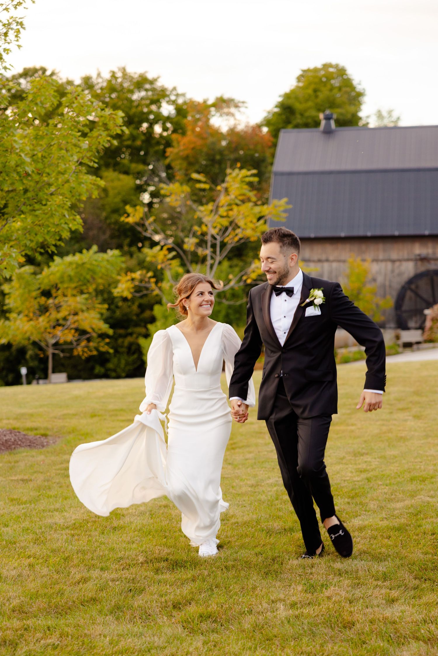 A couple in wedding attire playfully running on a green lawn with a barn in the background.