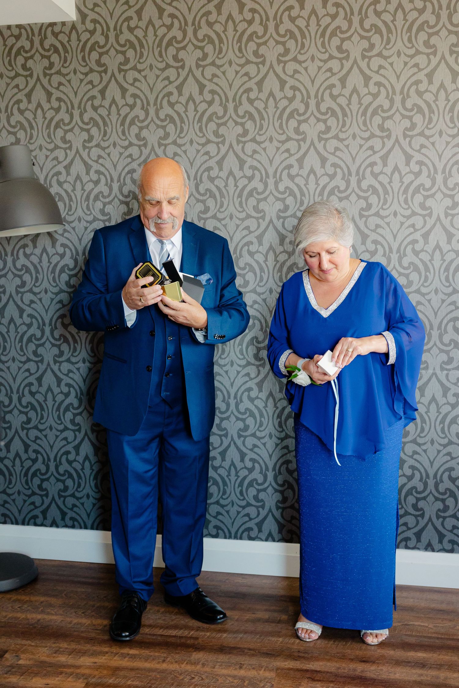 Two people in royal blue attire examine jewelry boxes against a grey patterned wall.