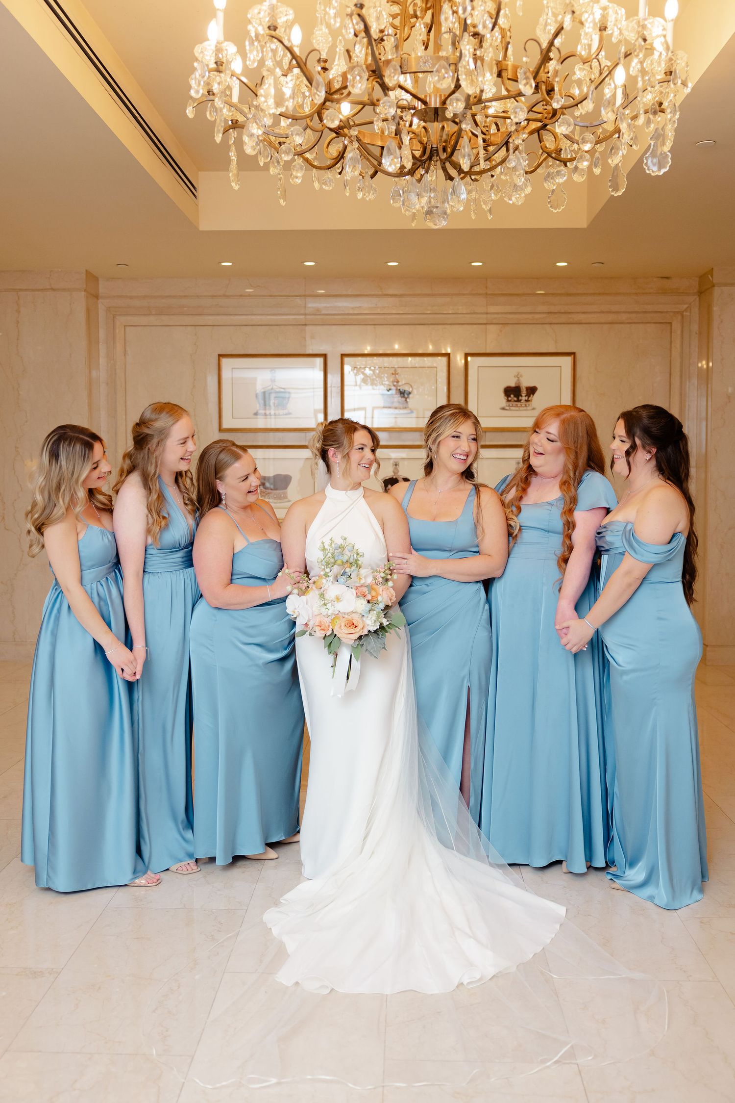 A bridal party in light blue bridesmaid dresses and white wedding gown pose creatively in an elegant room with chandeliers.