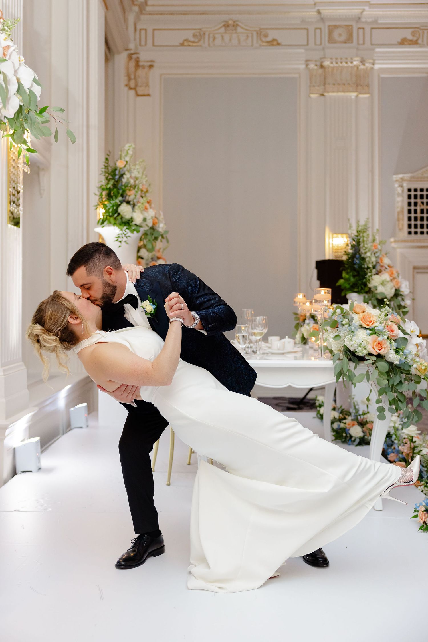 Romantic dip kiss sequence between newlyweds in an ornate white ballroom with floral decorations.