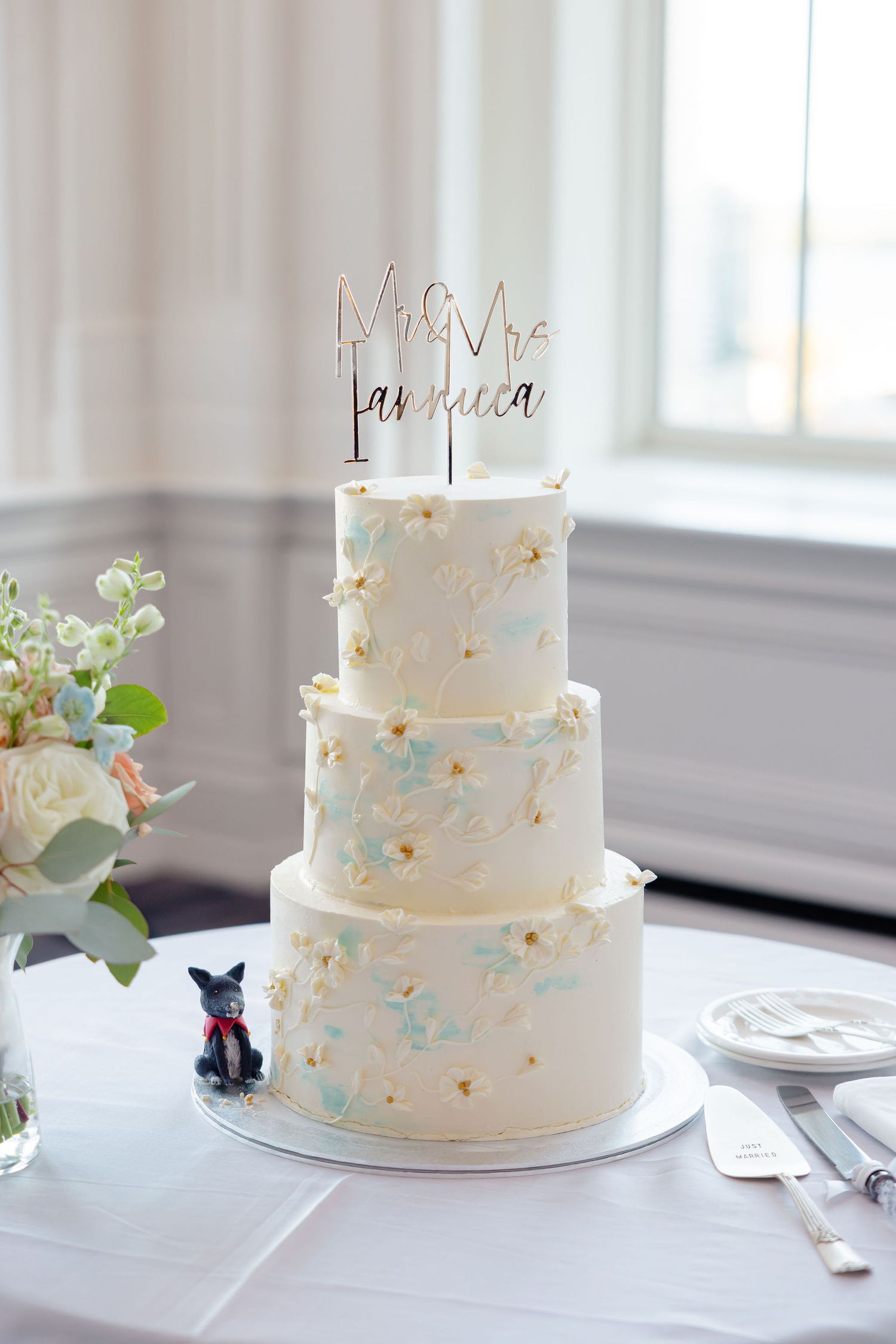 Three-tiered white wedding cake with delicate blue accents and monogram topper on white tablecloth.