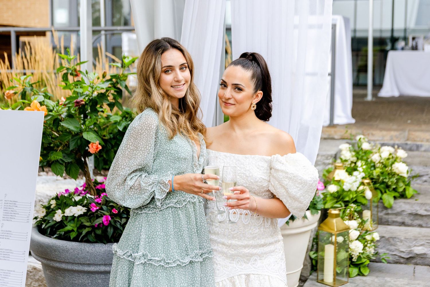 Two guests in elegant dresses enjoy drinks at an outdoor wedding reception with floral decorations.