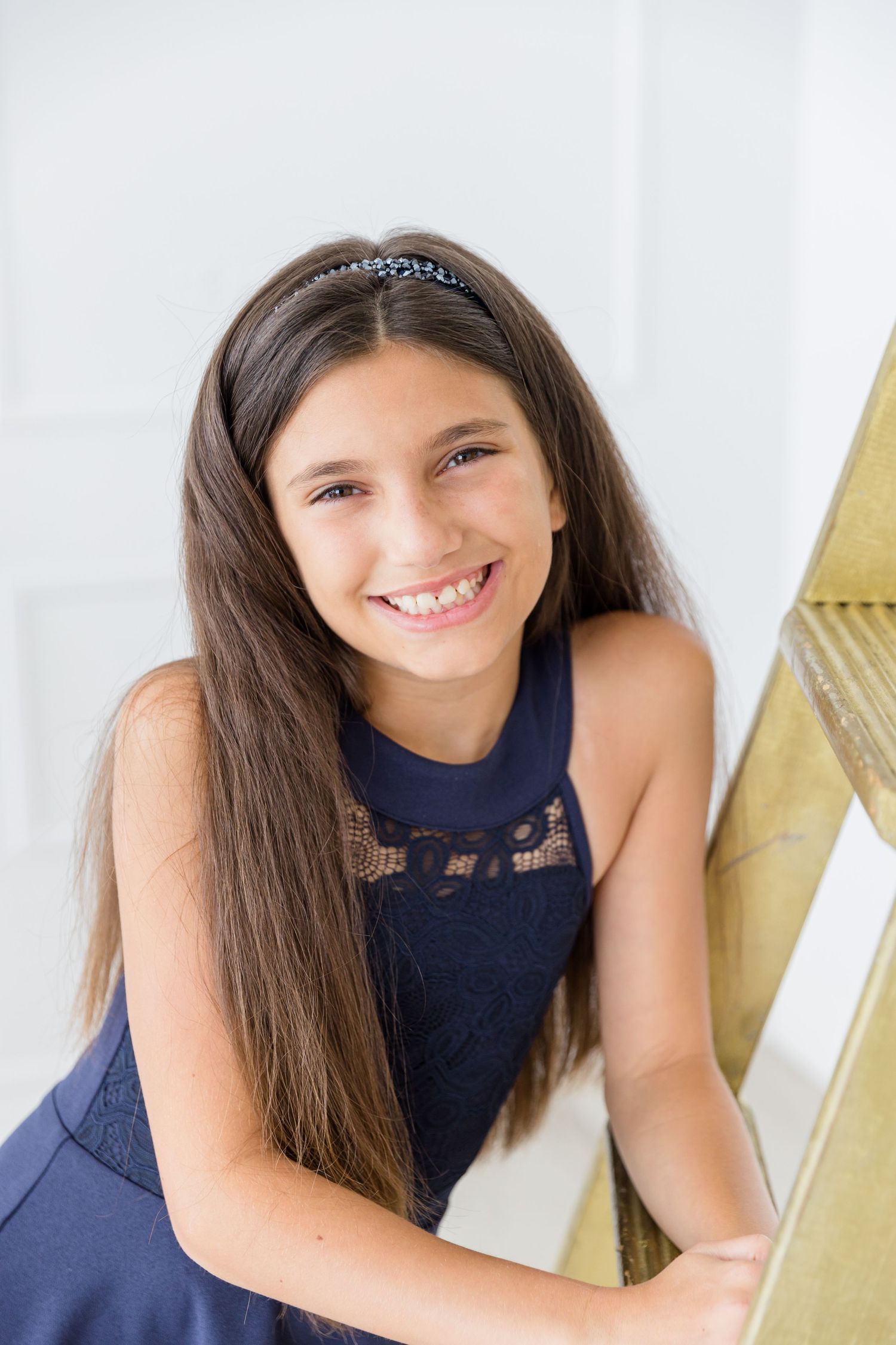 Young person wearing navy blue lace dress smiles while posing against white wall with gold accent.