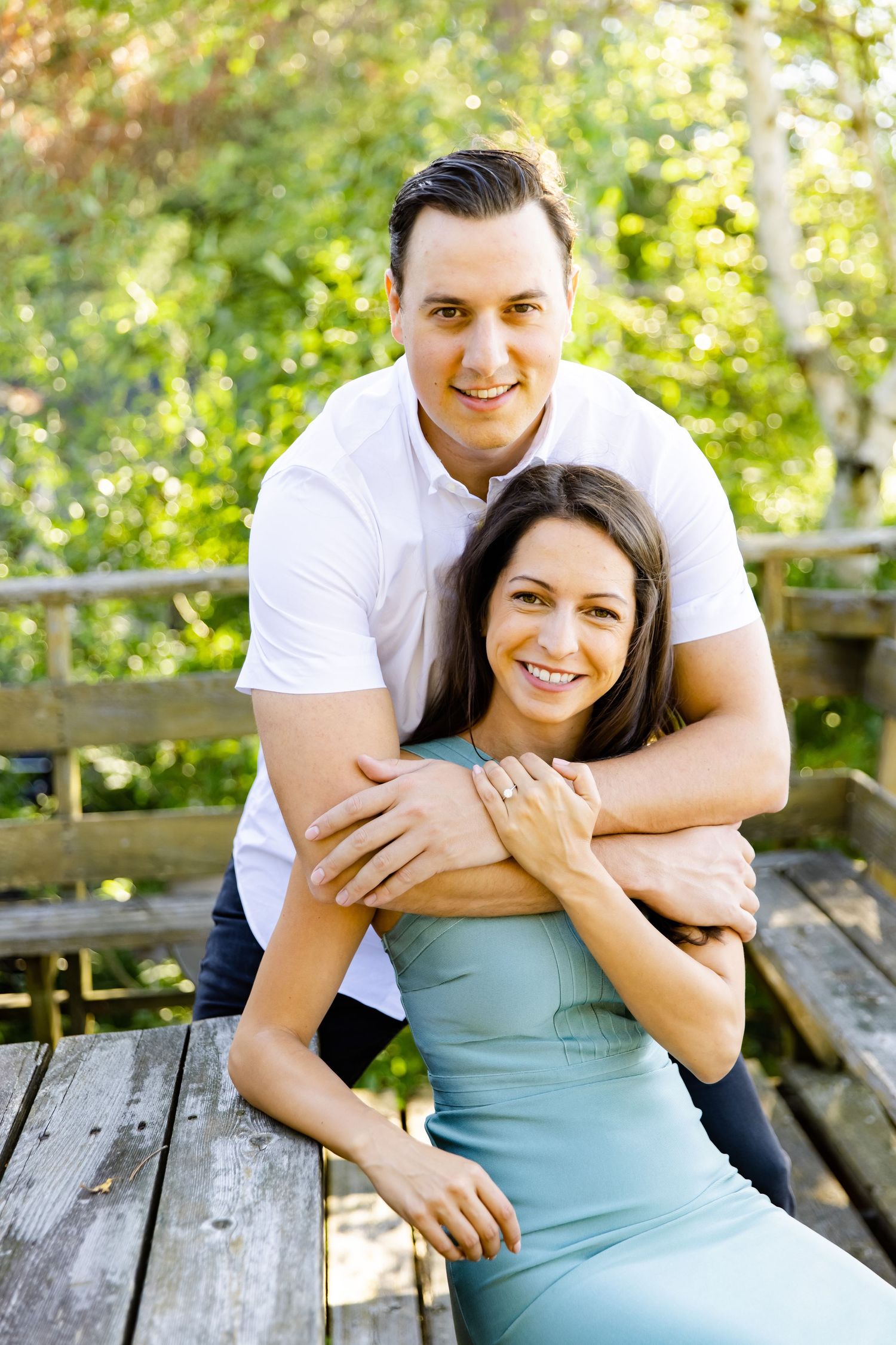 Couple shares tender moment on rustic wooden bench surrounded by summer foliage.