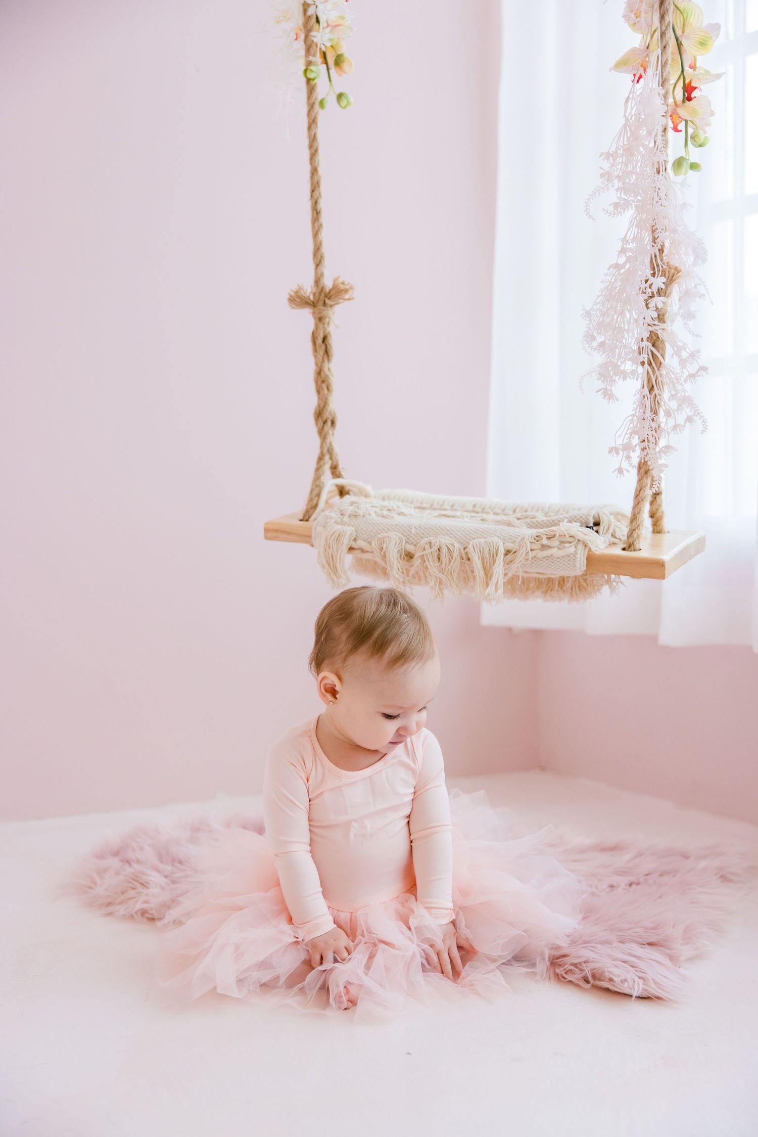 Baby in pink tulle dress sitting near a macrame swing in a soft pink room.