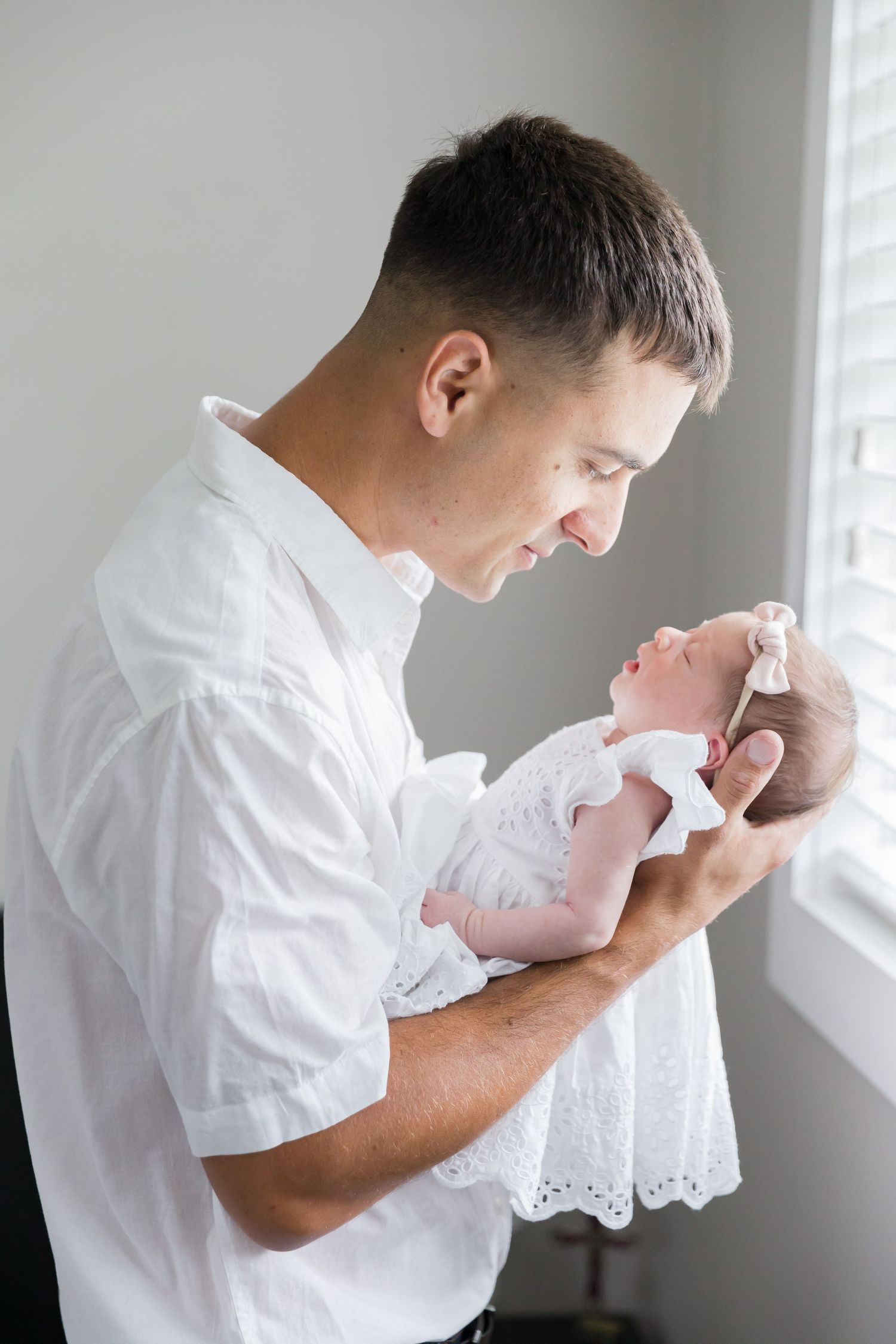 Parent gazes lovingly at newborn baby wearing delicate white dress next to bright window.