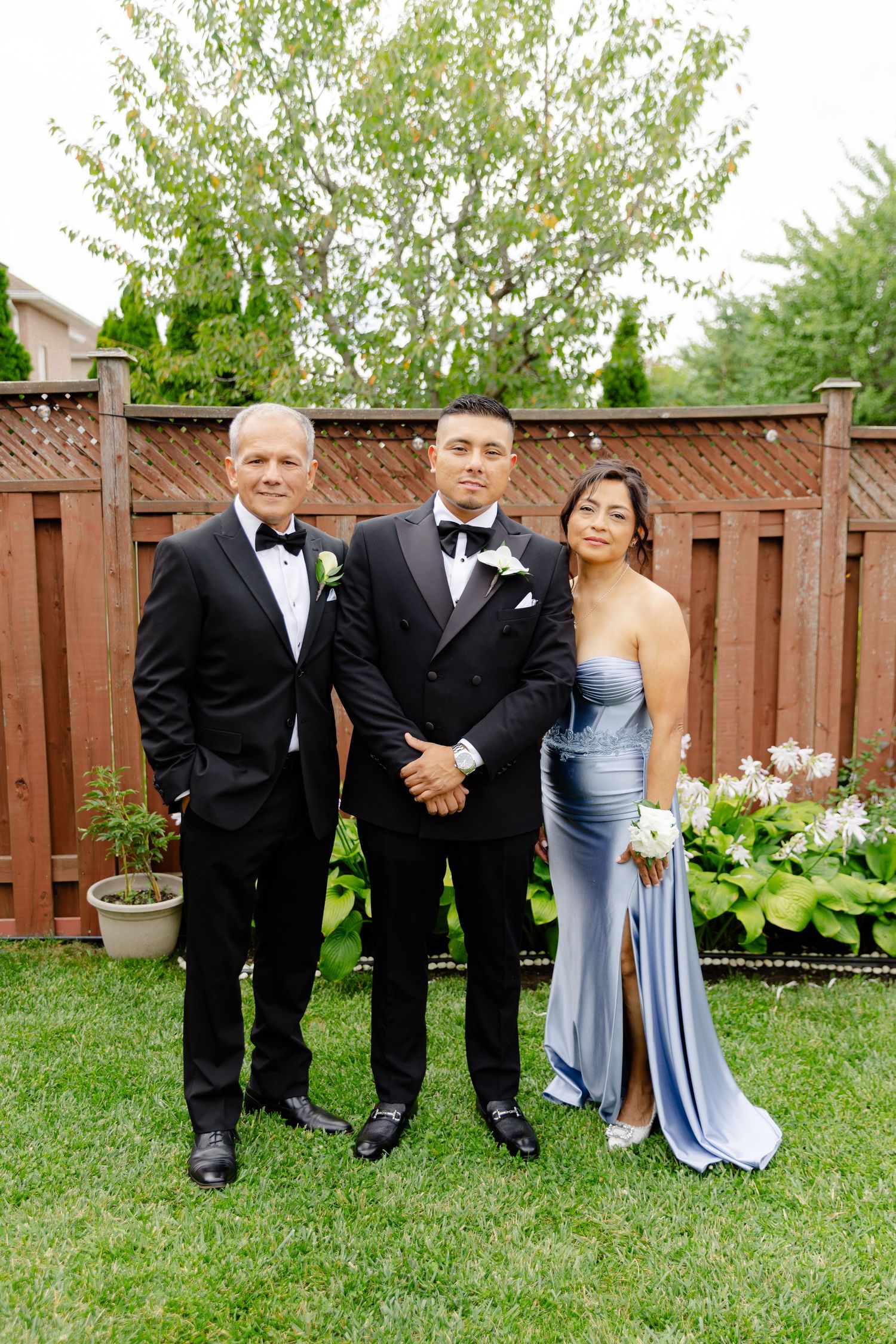 A formal wedding party in black tuxedos and blue dresses poses together on a green lawn with a wooden fence backdrop.