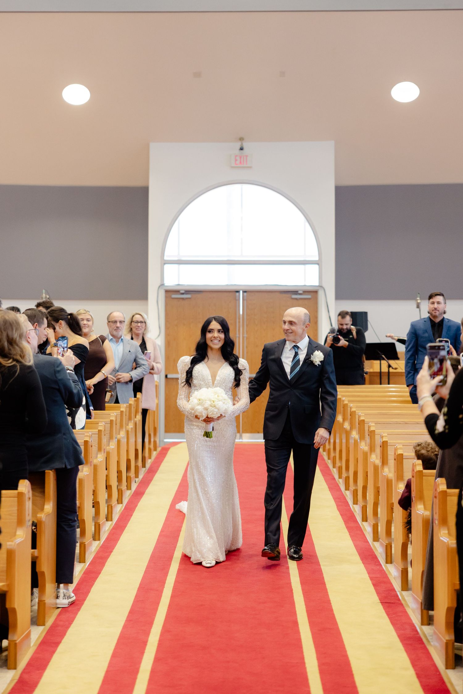 Wedding couple walks down red carpeted church aisle while guests take photos.