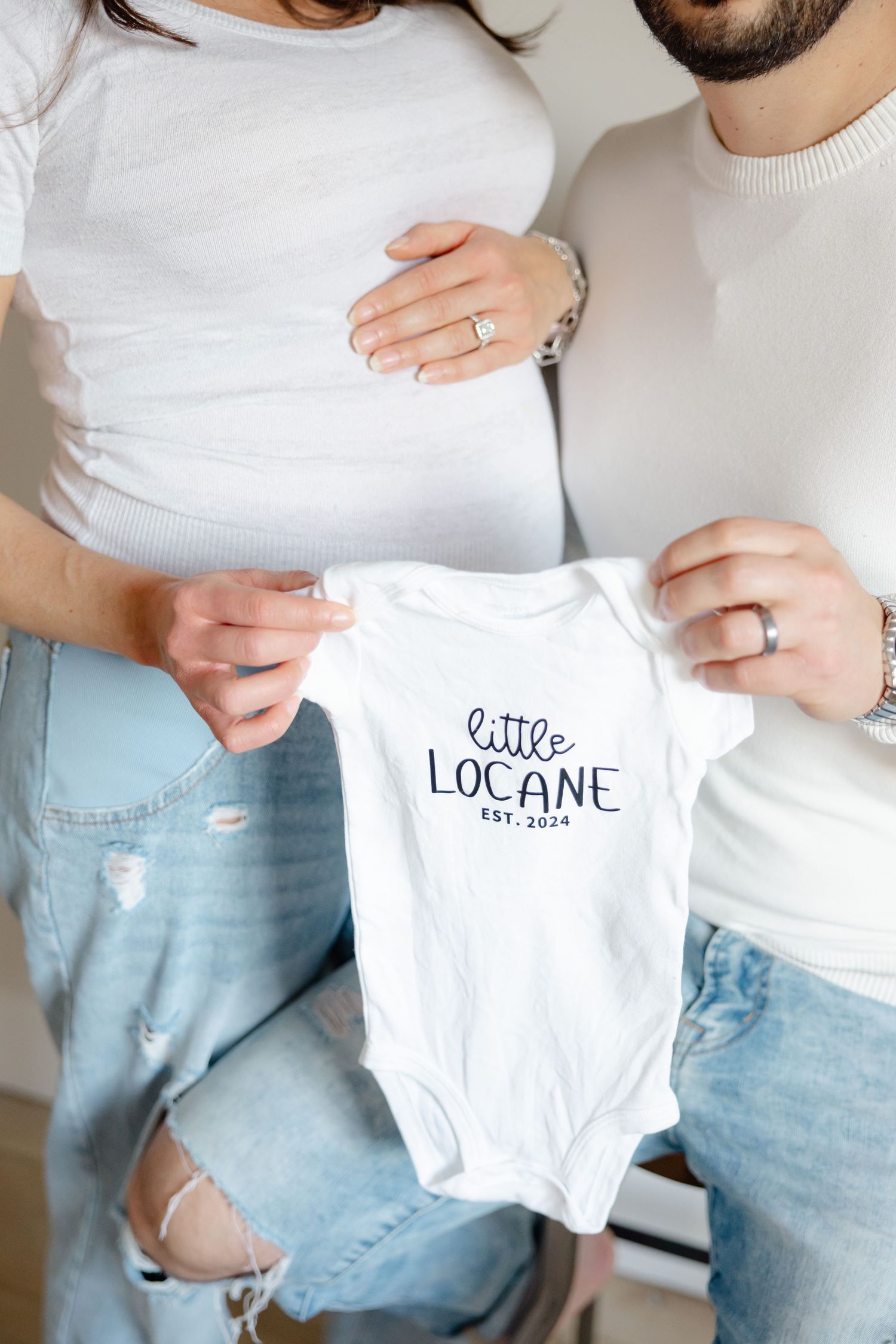 A couple holds up a baby onesie with 'Little Locane' printed on it.