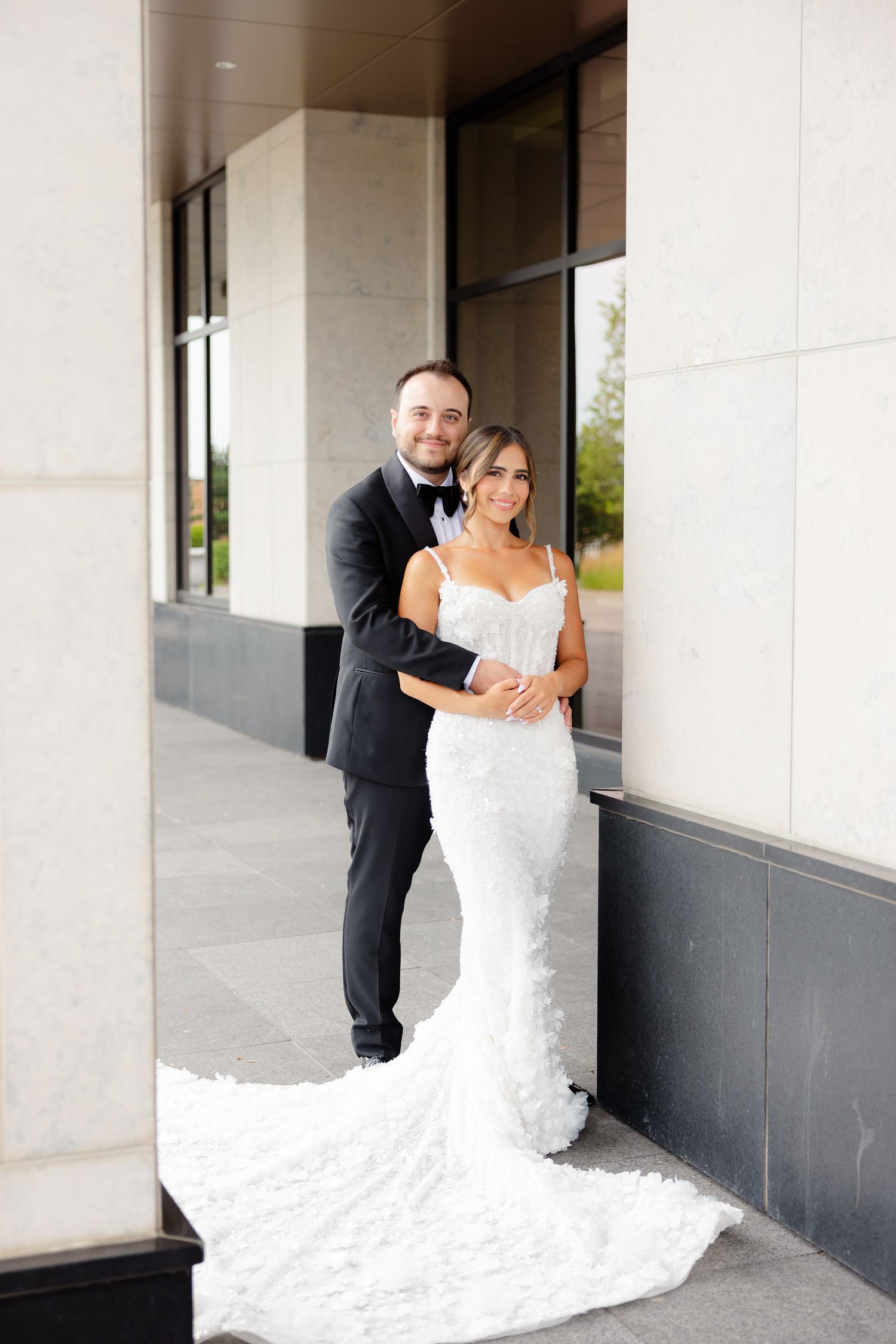 A couple in formal wedding attire shares playful poses in front of a modern building exterior.