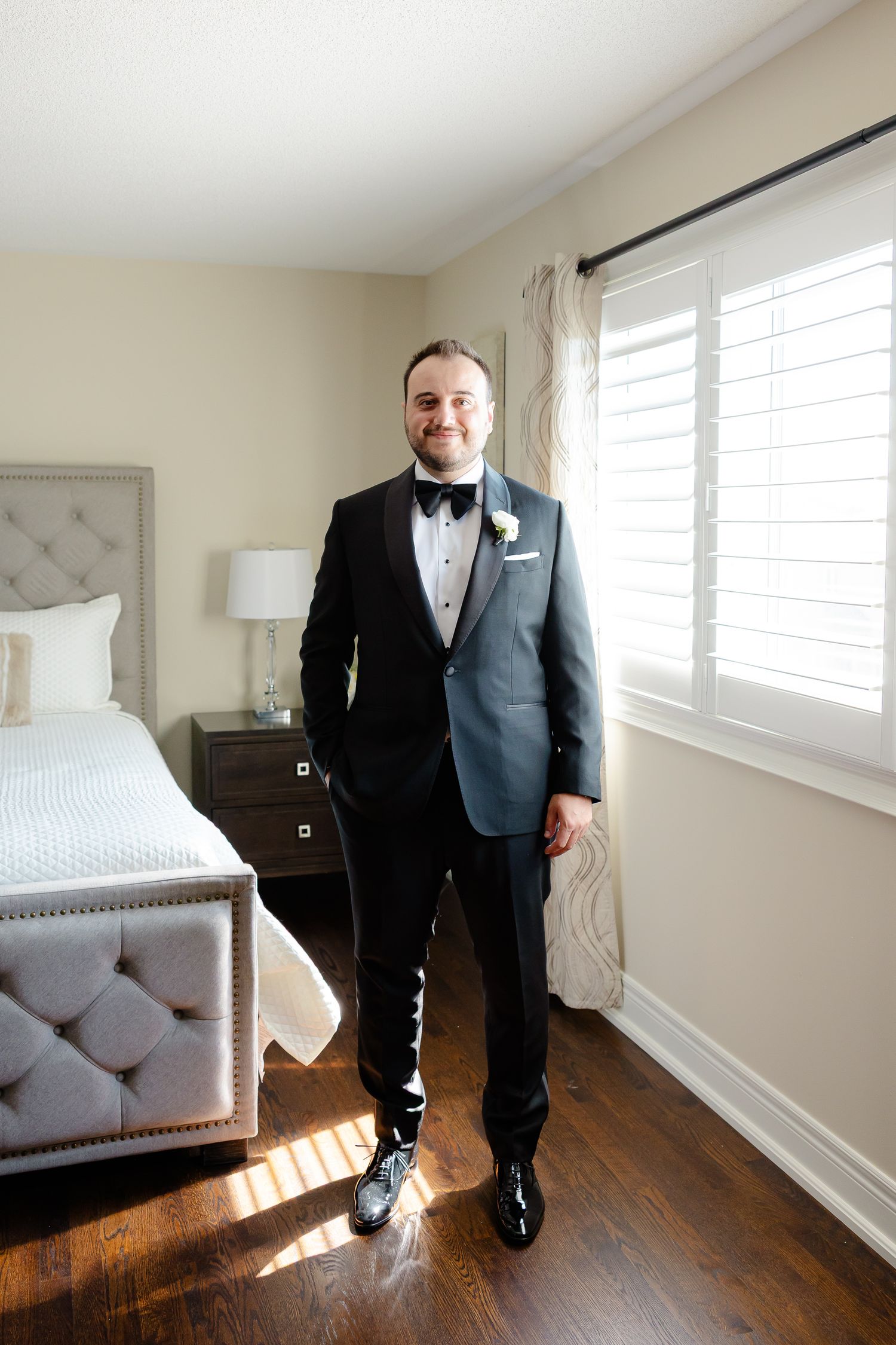 A person wearing a formal black tuxedo and bowtie stands in a sunlit bedroom with hardwood floors and white decor.