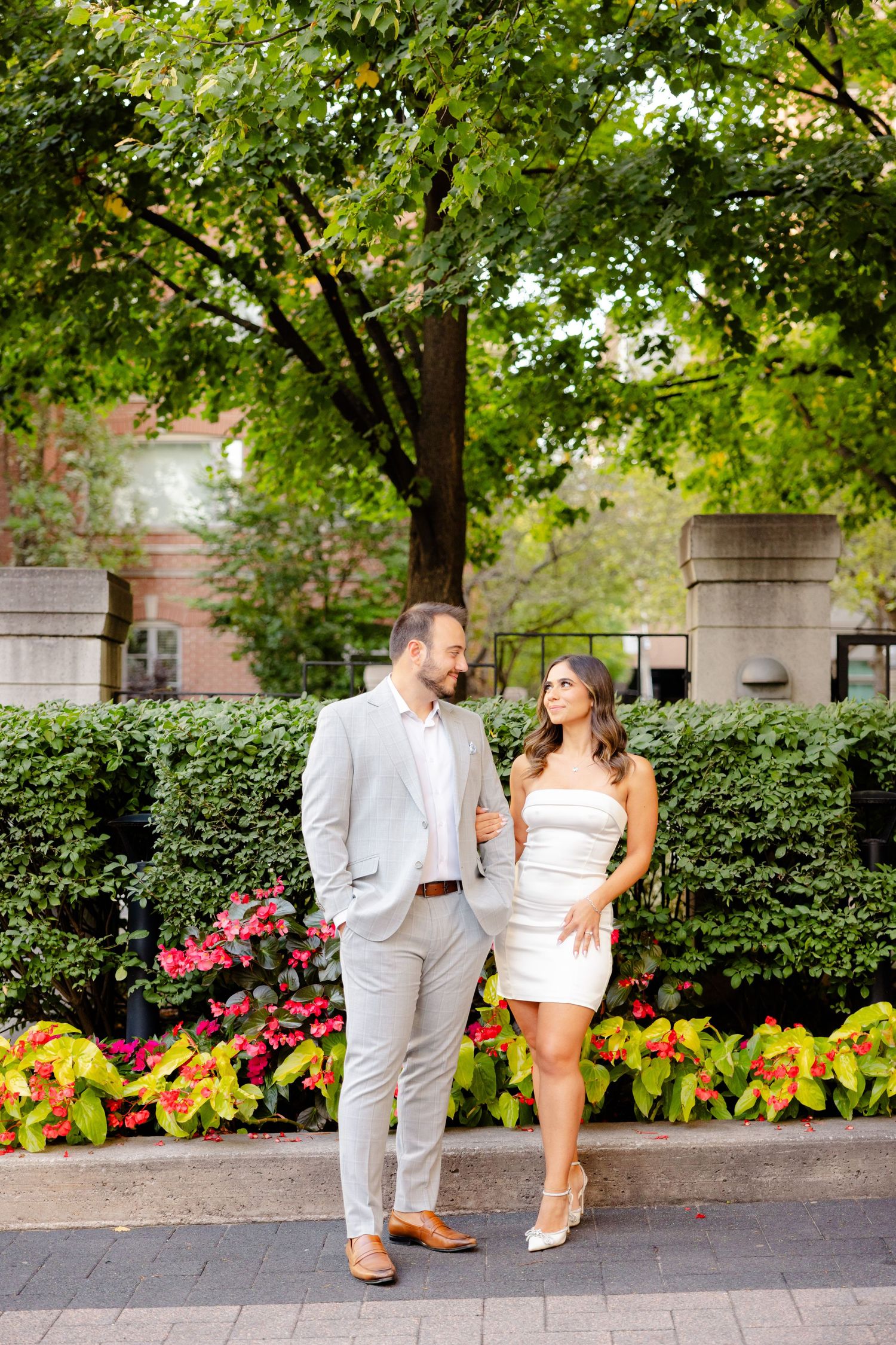 A couple shares a romantic moment walking along a garden path with trees and flowers in the background.