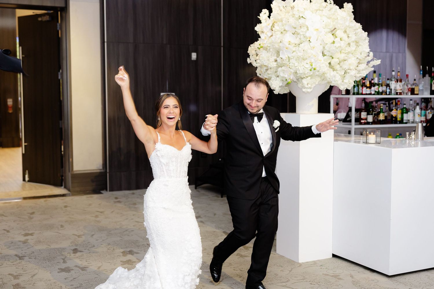 Joyful newlyweds dance together at their wedding reception next to an elegant white floral arrangement and bar setup.