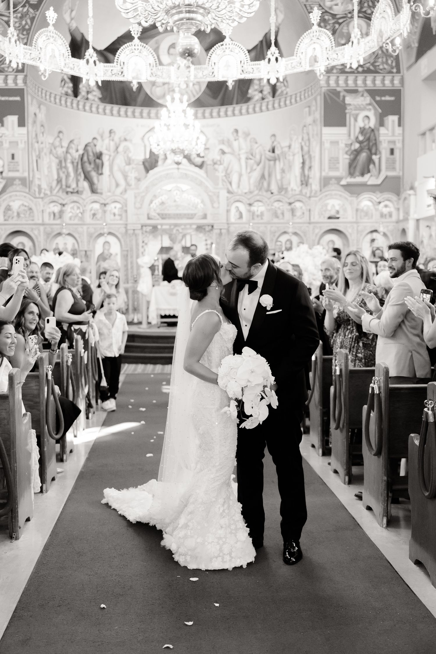 Wedding ceremony in progress inside an ornate church with guests seated in wooden pews and decorative murals on walls.