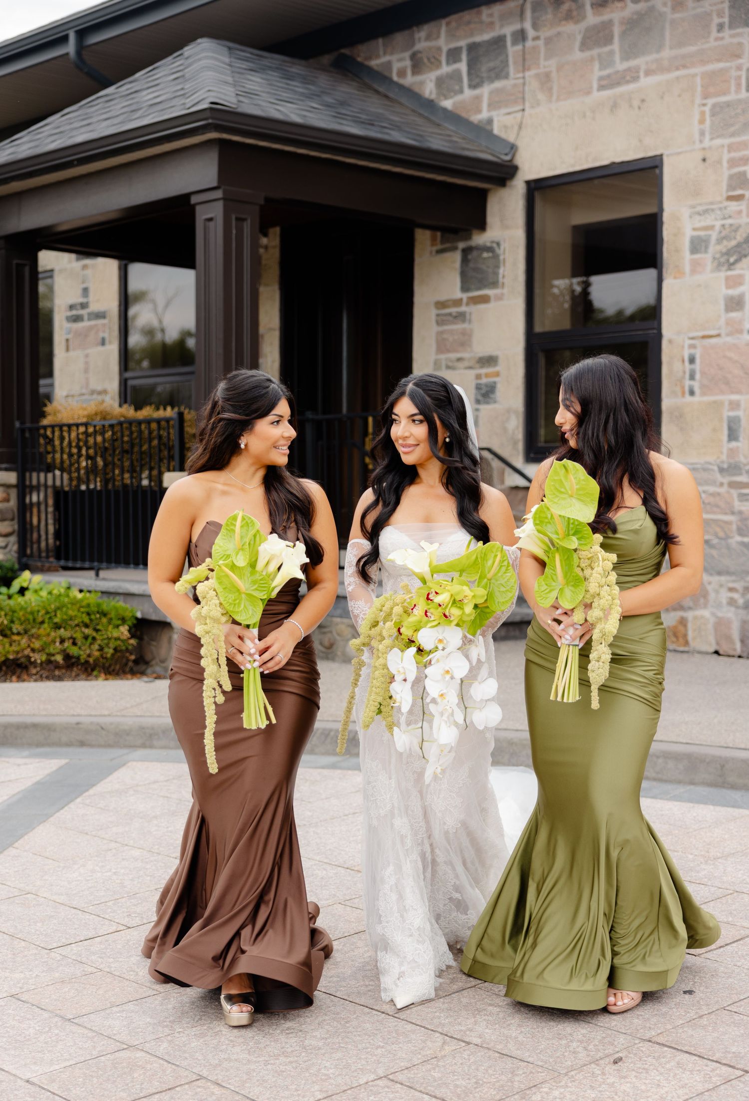 Bridal party in brown and olive dresses posing with bouquets outside stone building entrance.