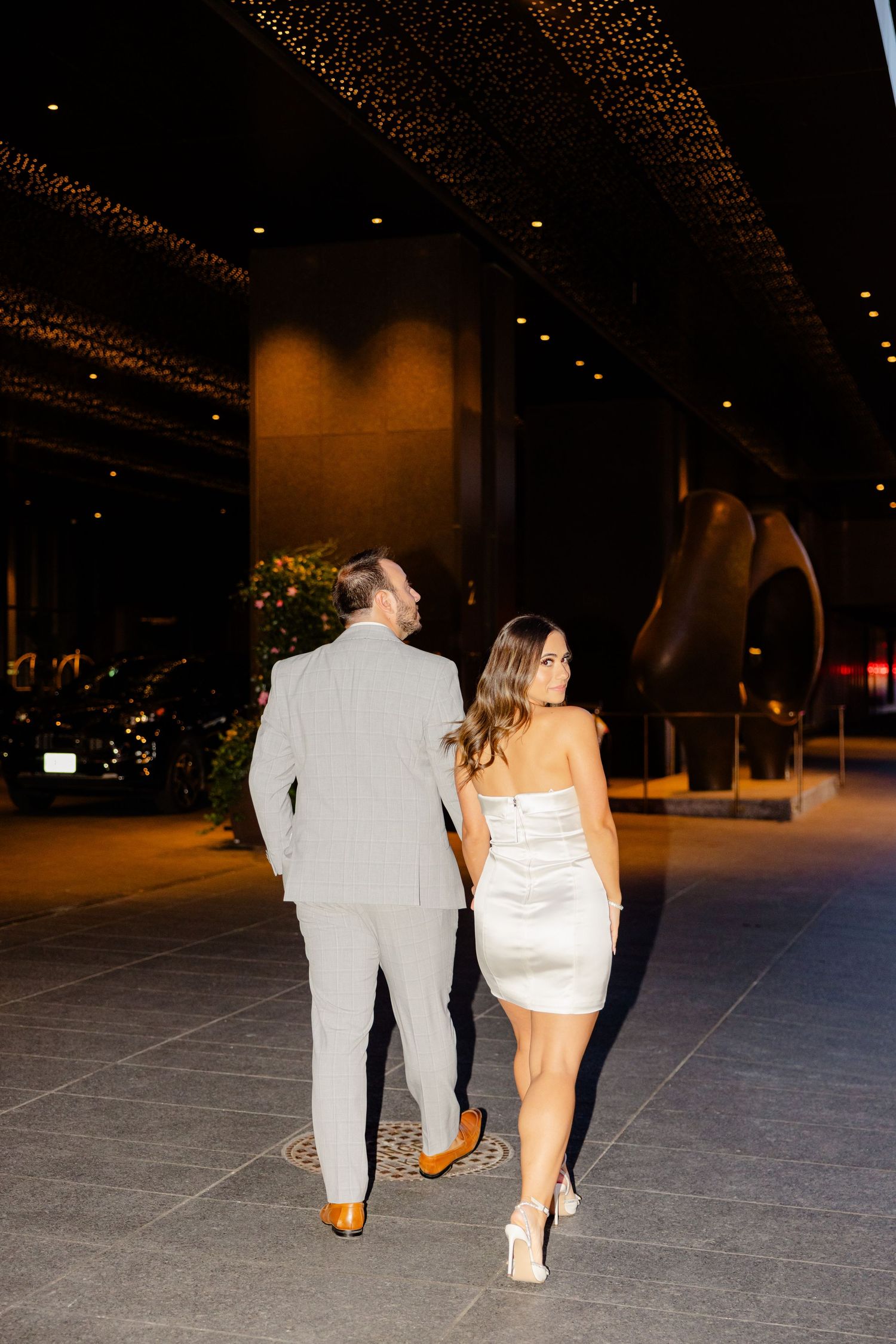 Couple walking away from camera, wearing a light gray suit and white mini dress in an evening setting.