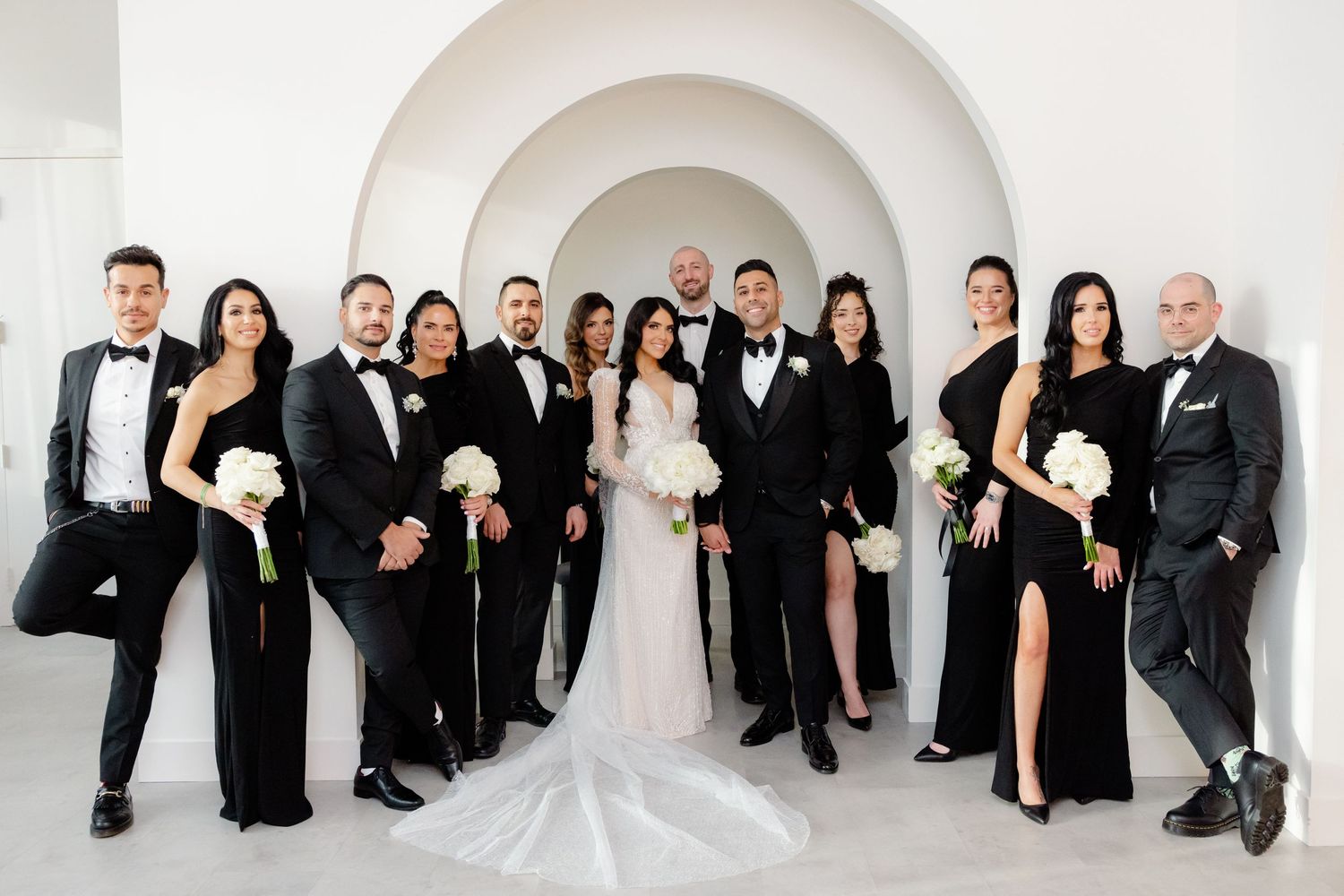 Wedding party in formal black attire and white gowns pose together for elegant group photo.