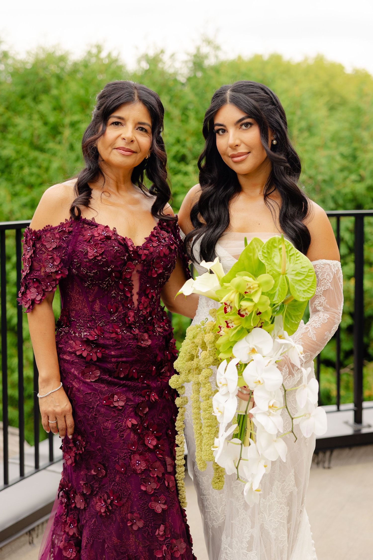 Two women in burgundy and white formal dresses pose together on an outdoor balcony during a special occasion.