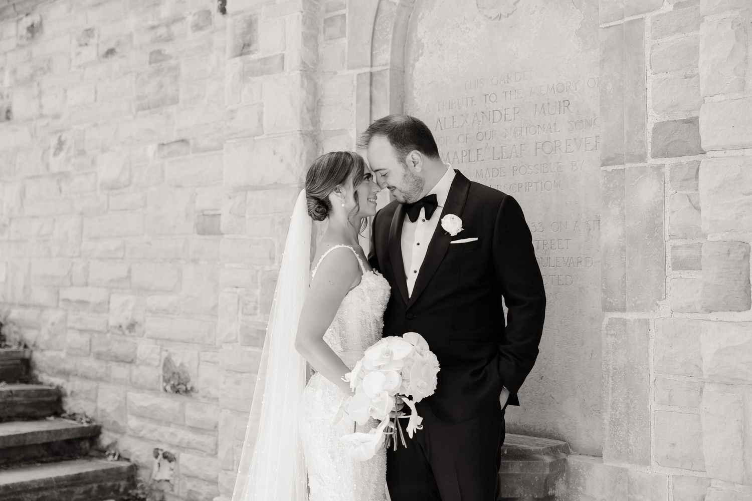 A black and white wedding photograph captured against a stone wall exterior of a church.