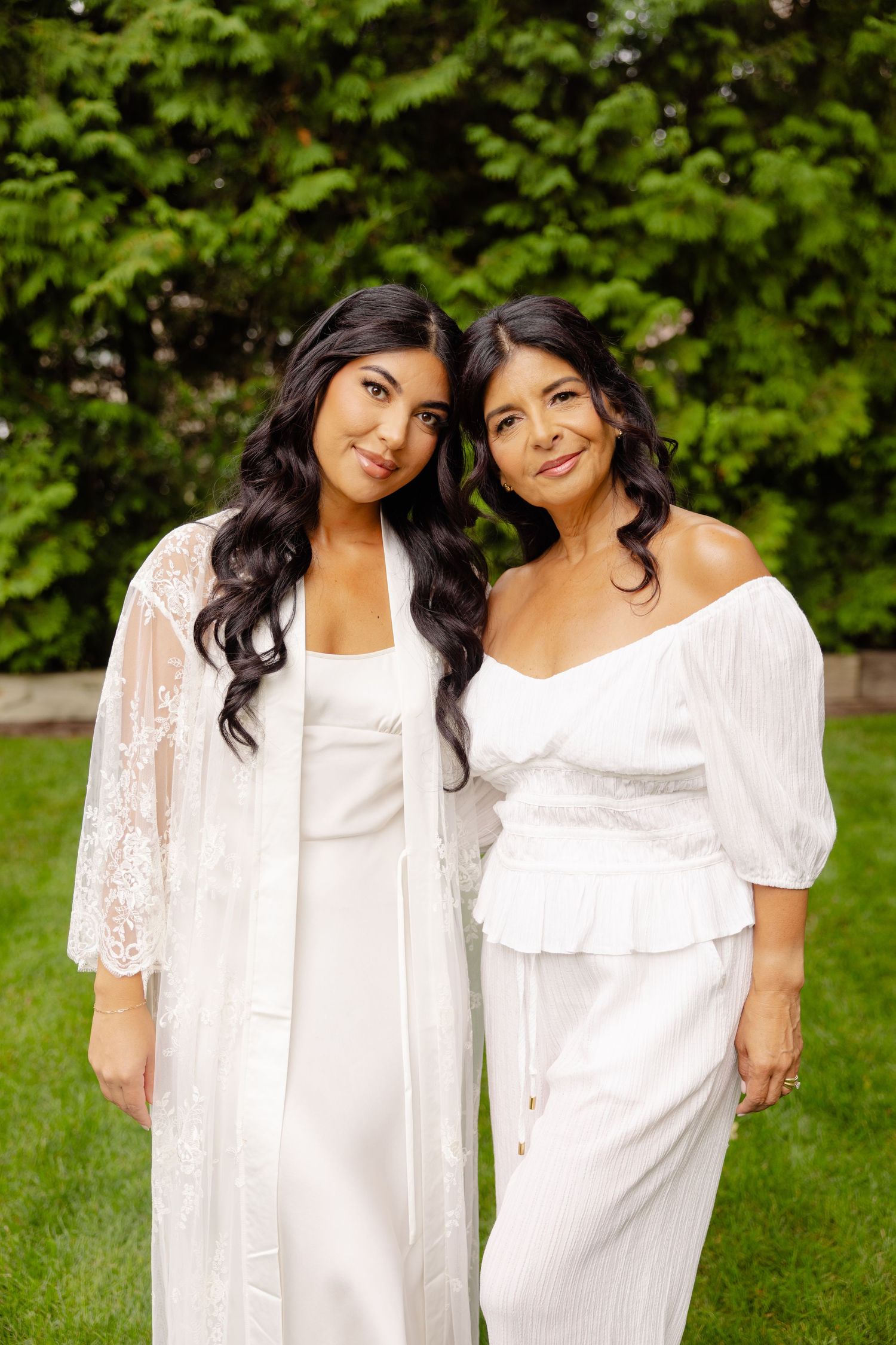 Two people wearing white outfits standing together on grass with green foliage in the background.