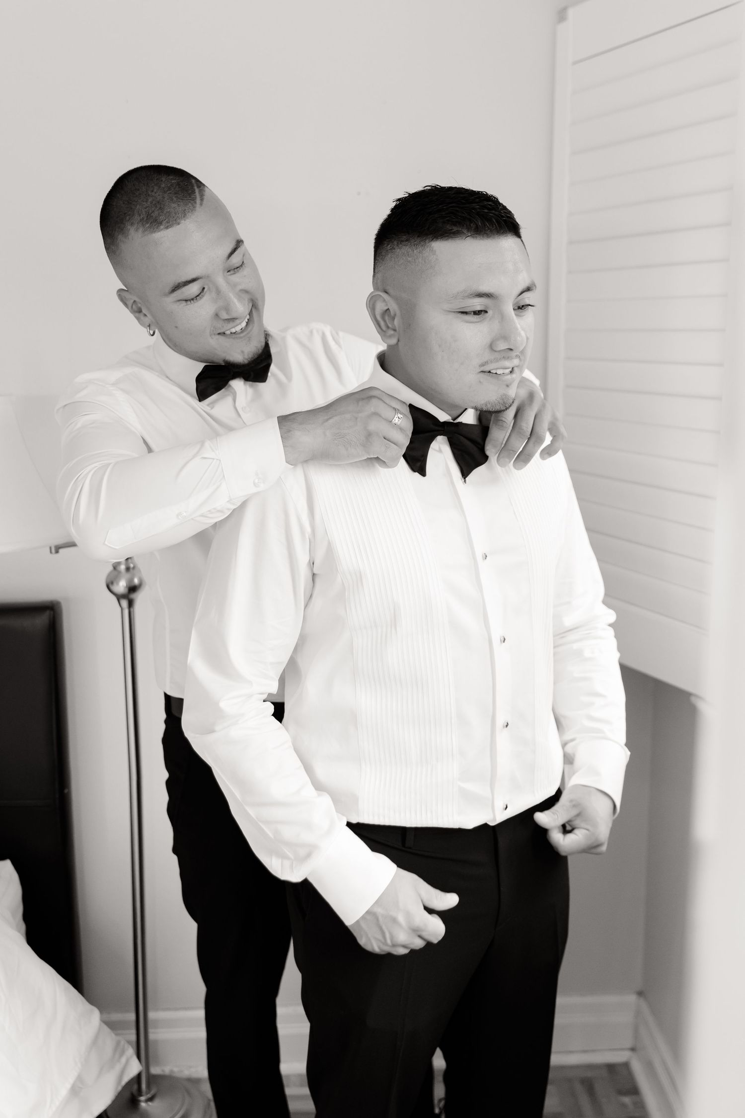 Two people in matching tuxedos laugh and embrace during a candid moment in a bright room.